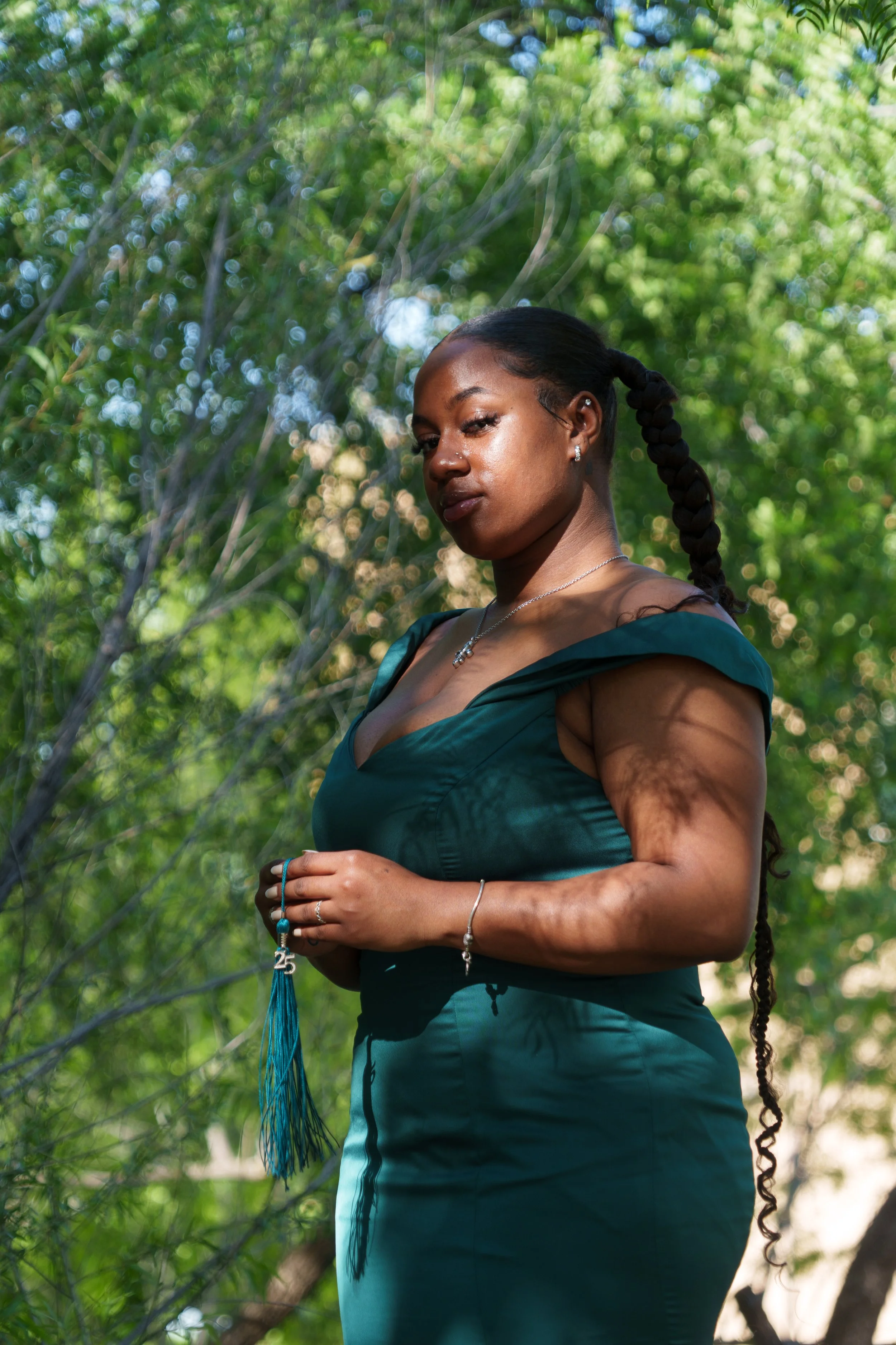 Portrait of a woman with braided hair standing outdoors among green trees, wearing a teal dress, holding a beaded tassel, and jewelry, with sunlight filtering through the leaves.