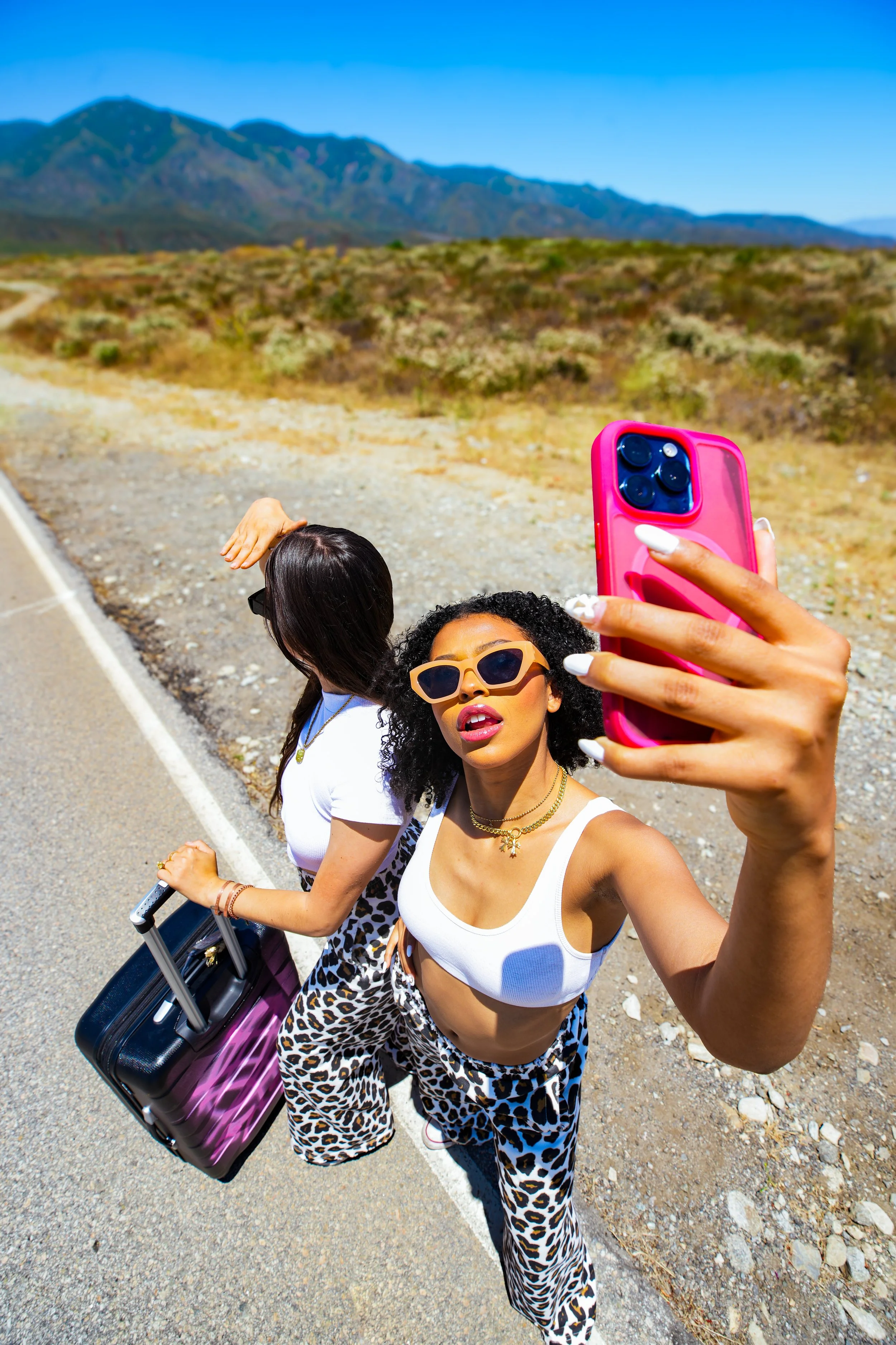 Two women taking a selfie on the side of a road with mountains in the background. One woman holds a pink phone, wearing sunglasses, a white crop top, and leopard print pants. The other woman carries a suitcase and wears a white t-shirt and similar le