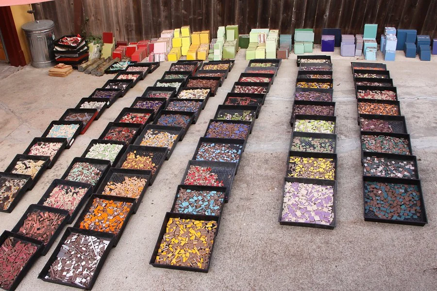 Multiple trays of colorful mosaic tiles arranged in rows on concrete ground, with stacks of boxes in various colors in the background and a wooden fence.
