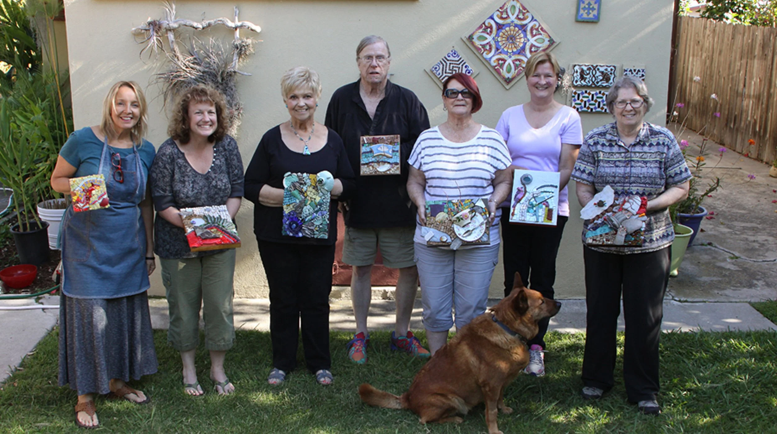 A group of eight women standing in a backyard garden, holding fabric or art projects, with a brown dog sitting in front of them. The background features decorative garden art on the wall, potted plants, and a wooden fence.