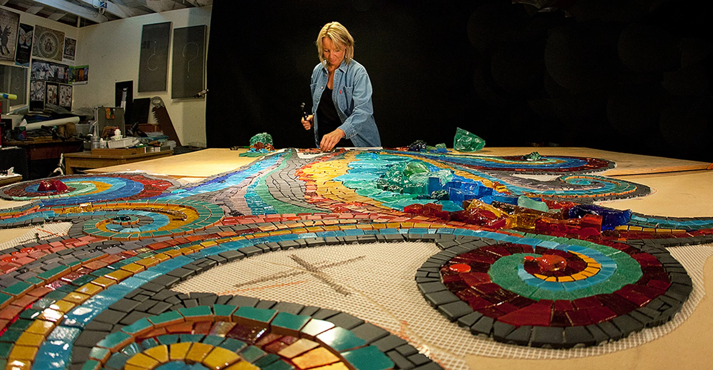 A woman working on a large colorful mosaic artwork on a table in an art studio.