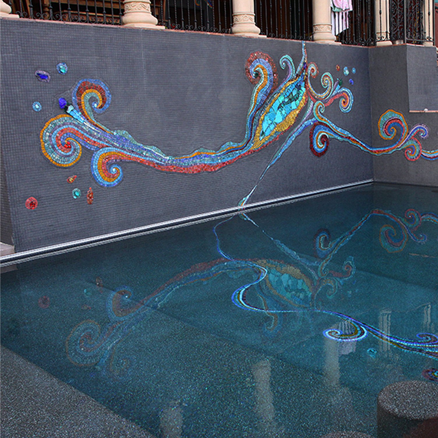 Colorful mosaic artwork on a dark tiled wall reflecting in a swimming pool below.