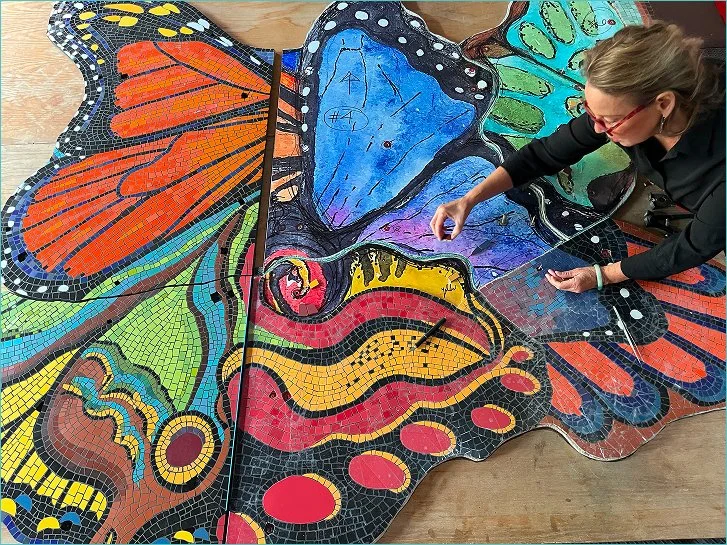 A woman working on a colorful butterfly mosaic art piece on a wooden surface.