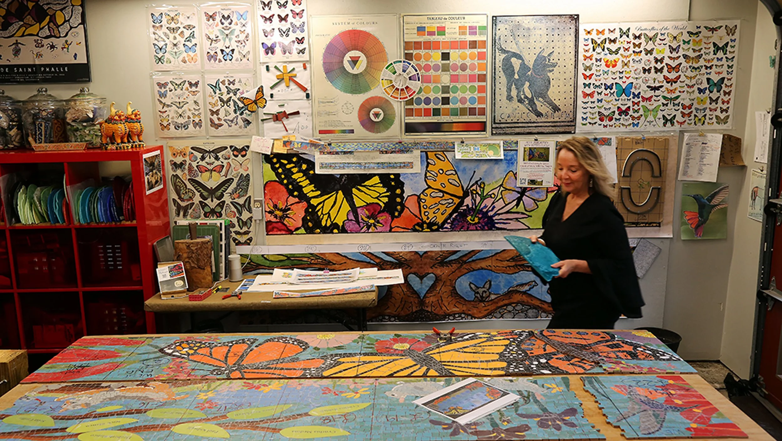 An art studio with tables displaying colorful butterfly-themed artwork, charts, and color wheels on the wall, a red shelving unit with jars and decorations, and a woman holding a piece of artwork, smiling.