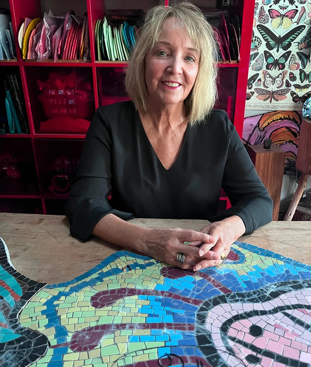 A woman with blonde hair and a black top sitting at a table with a colorful mosaic art piece. Behind her are pink shelves filled with various colored files and a display of butterfly posters.