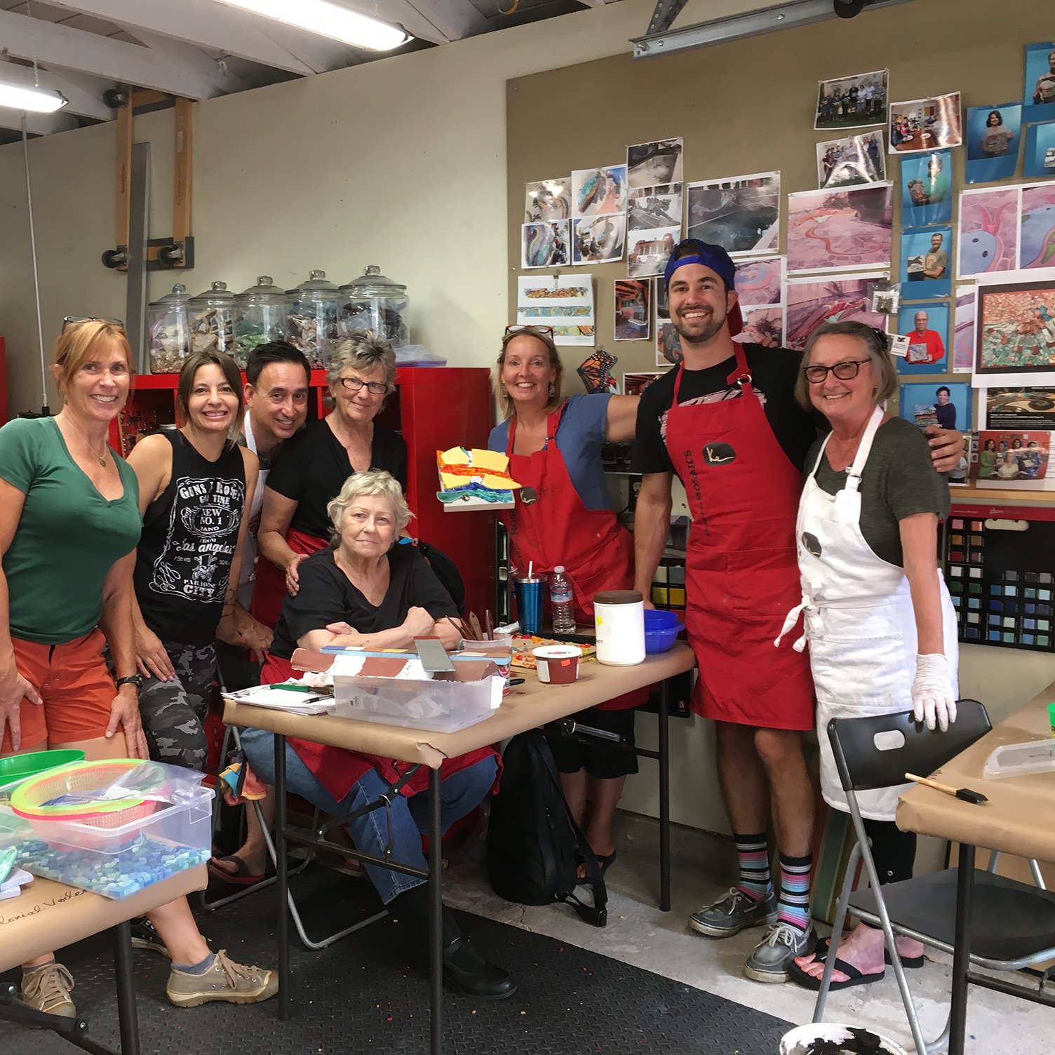 Group of eight people, some wearing aprons, gathered around a table with art supplies in a craft room, smiling at the camera.