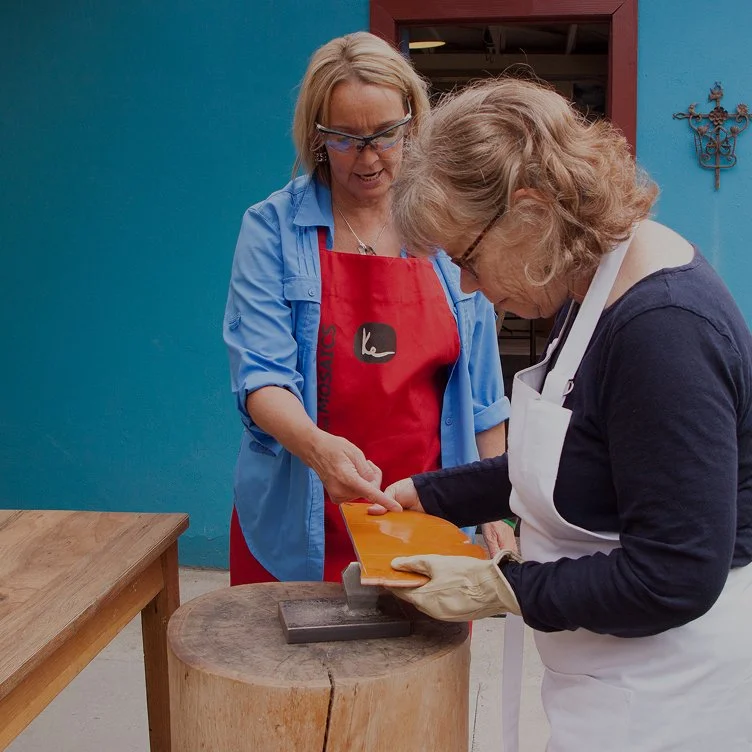 Two women, one wearing a blue shirt and red apron and the other in a white apron and gloves, are working together to carve a piece of orange soap on a wooden chopping block.