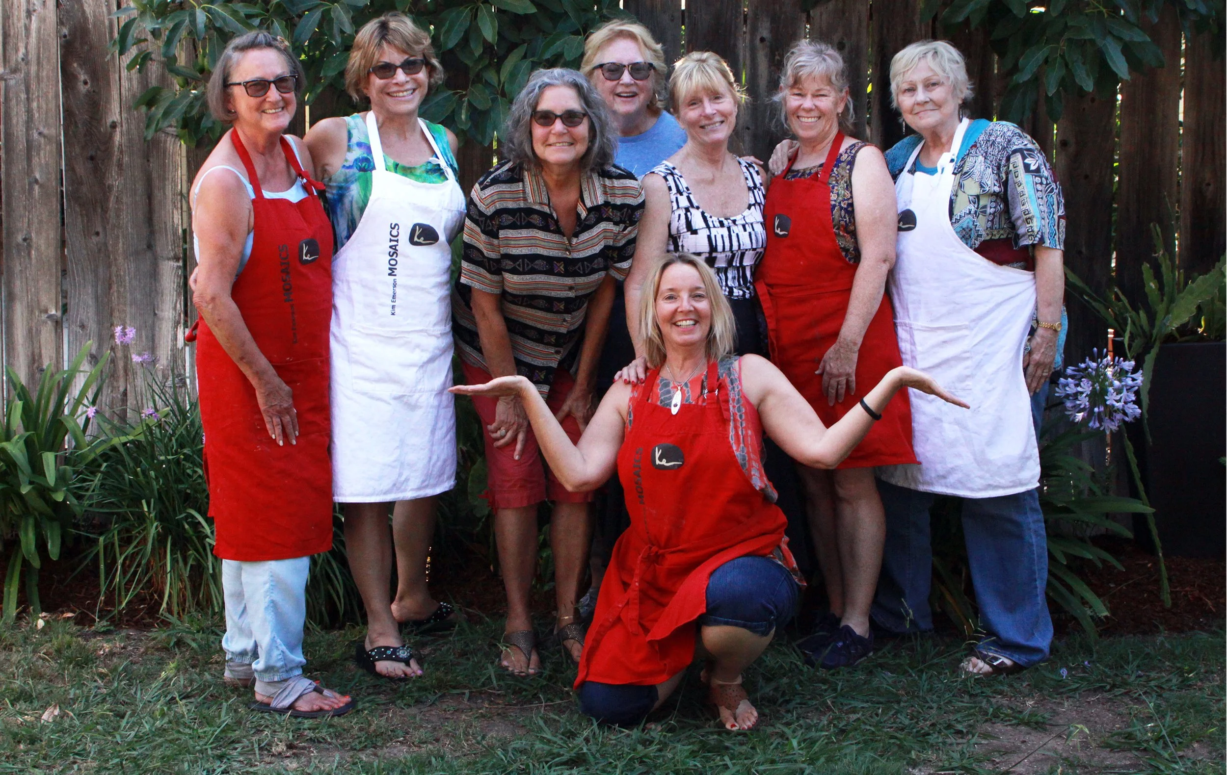A group of nine women, most wearing aprons, posing outdoors in front of a wooden fence and green foliage. They are smiling and some are wearing sunglasses.