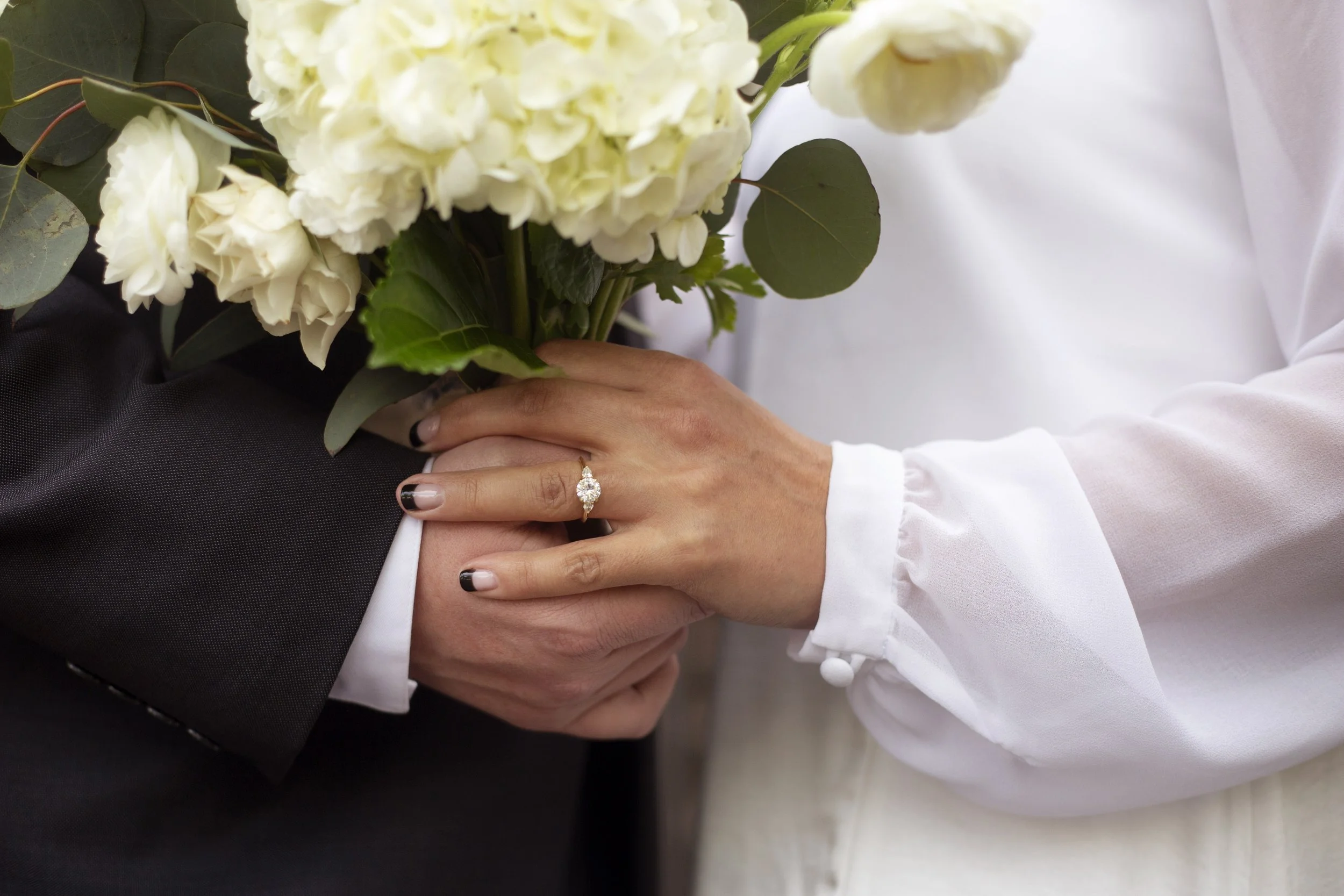 A close-up of a bride and groom holding hands, with the bride wearing a wedding ring and a white blouse and the groom in a black suit. The bride is holding a bouquet of white flowers.