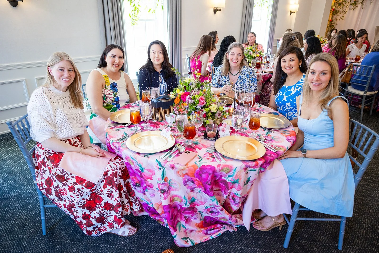 Group of six guests smiling at camera while seated at floral tablecloth table during Morrison House celebration event