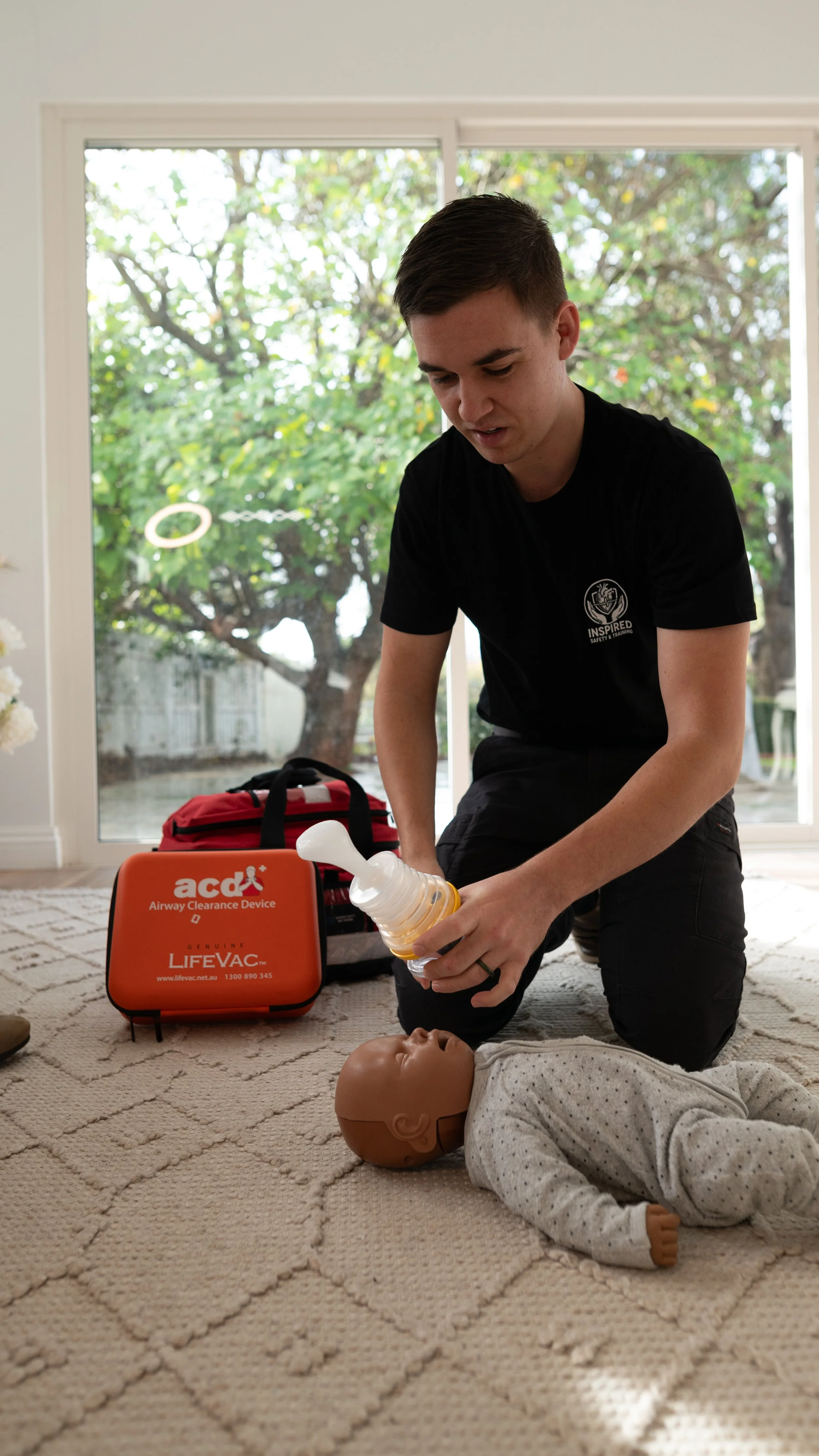 A man practicing CPR on a training mannequin with a baby doll face, in a room with large windows and green trees outside. CPR training equipment and a red emergency response bag are visible.
