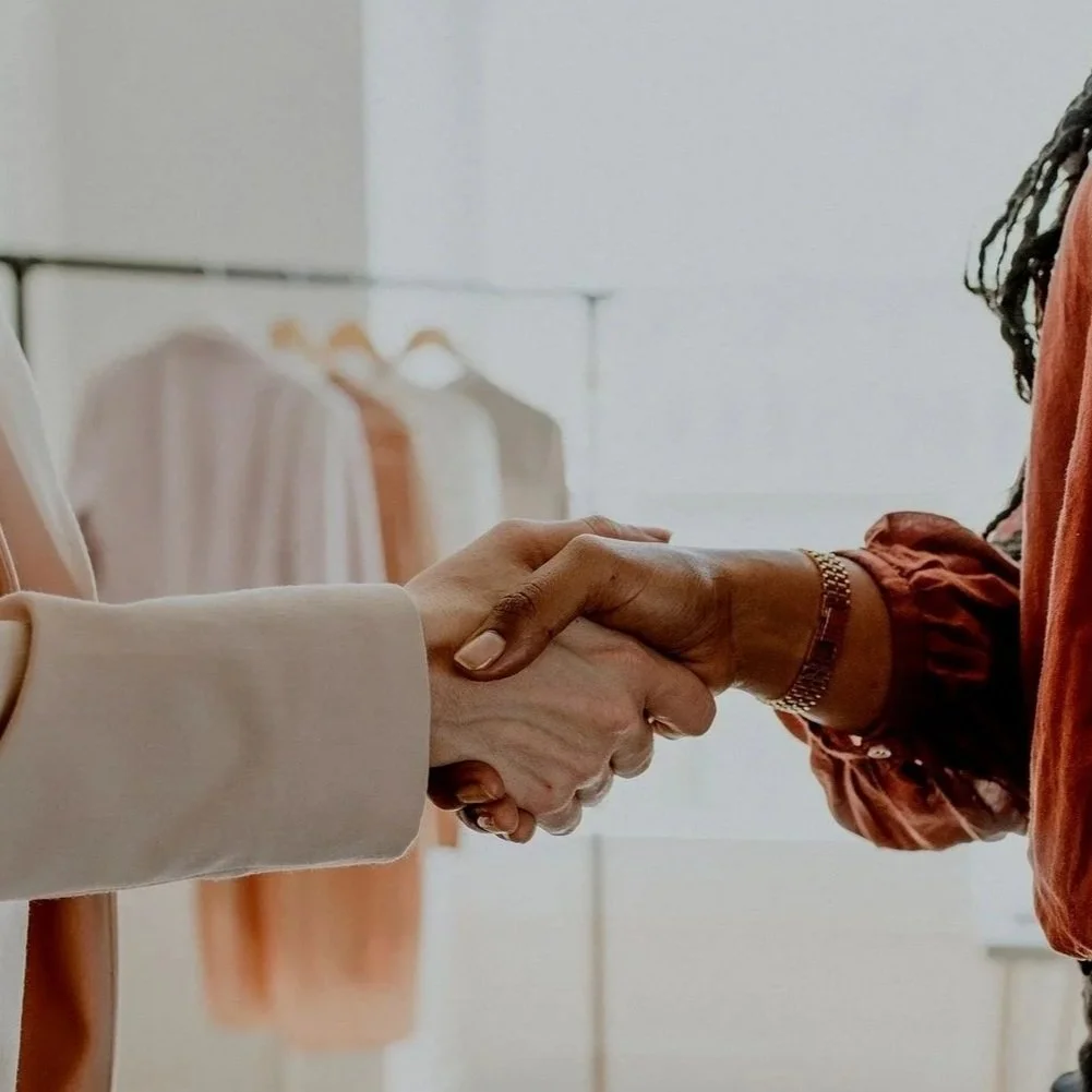 Two people shaking hands, one wearing a beige blazer and the other wearing a rust-colored top with a bracelet.