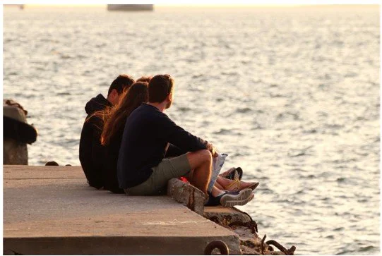 Three people sitting on a concrete pier by the water during sunset, facing the ocean.