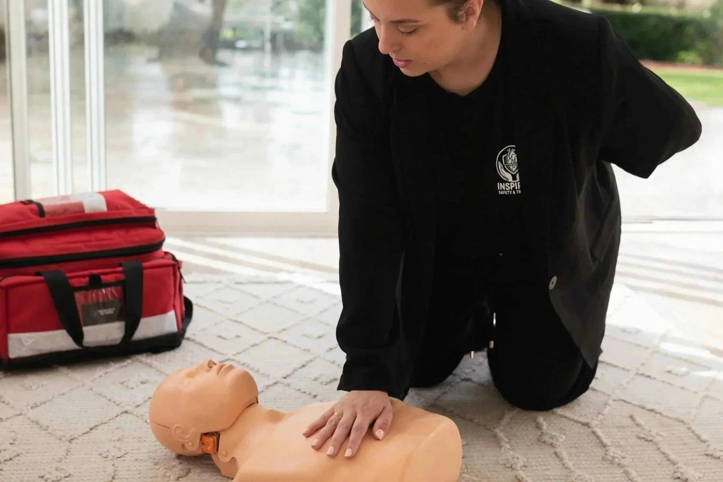 A person practicing CPR on a training dummy inside a building with a red emergency medical kit nearby.
