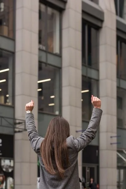 Back view of a woman with long brown hair raising her fists in triumph on a city street, with a modern building in the background.