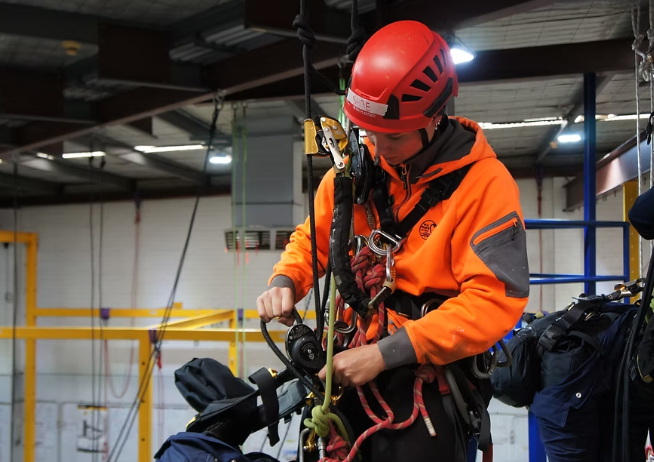 A person wearing an orange jacket, a red helmet, and safety harness preparing climbing or rescue gear indoors.