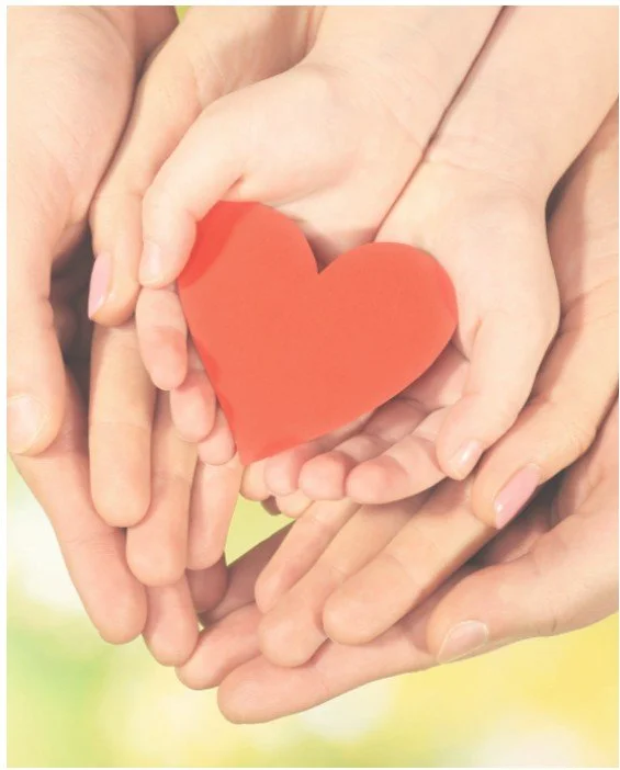 Multiple hands surrounding and holding a red heart-shaped object together.