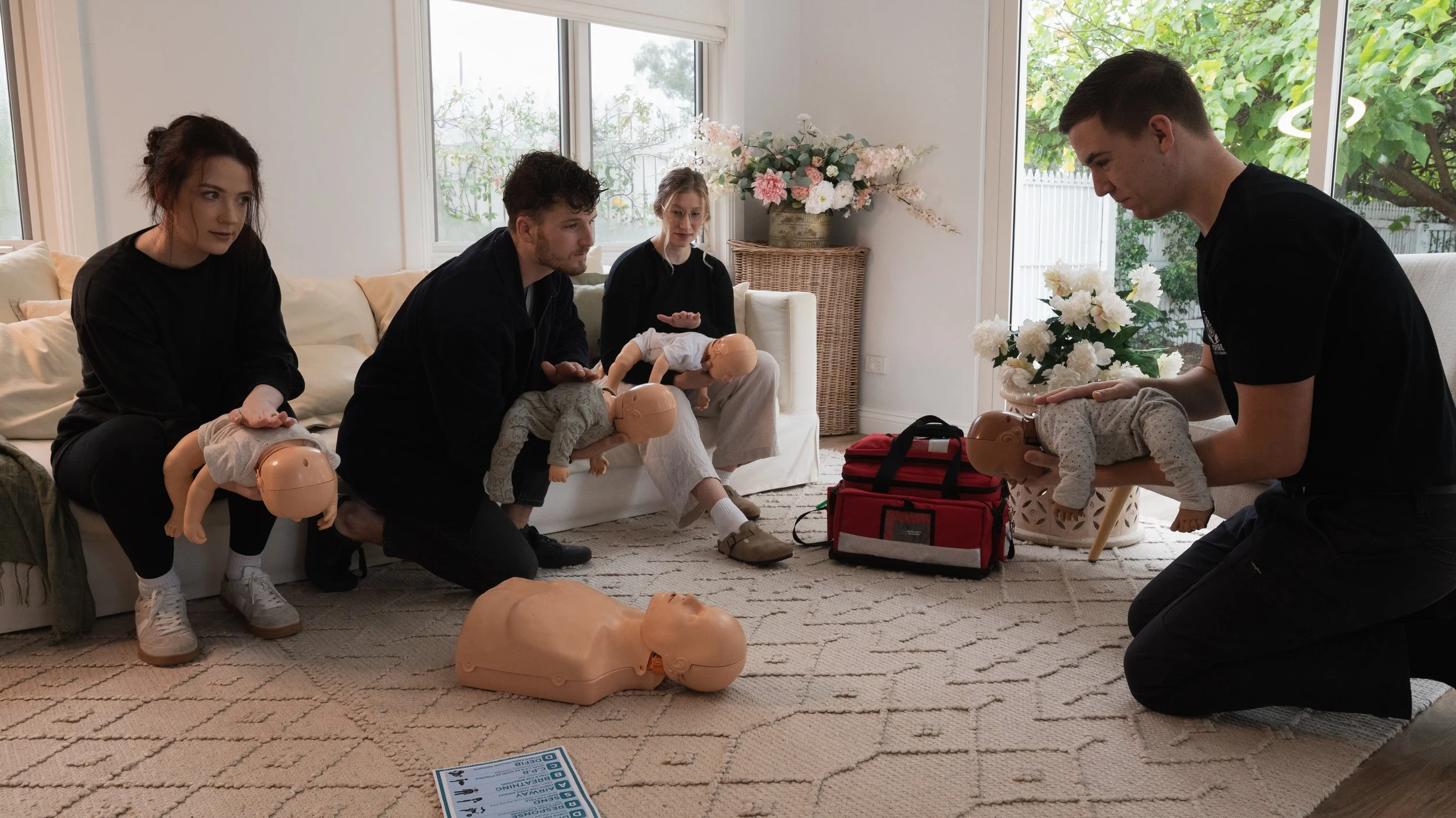 Group of five people participating in a CPR training session with mannequins and baby dolls in a living room decorated with flowers and large windows.