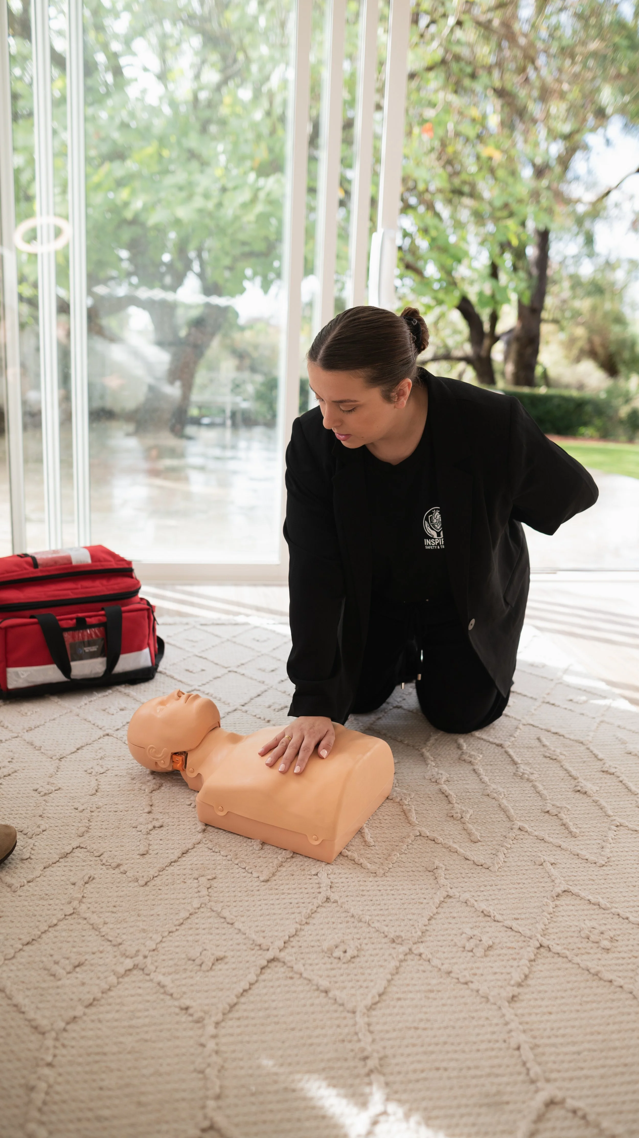 A woman practicing CPR on a training mannequin inside a room with large windows and green outdoor scenery.