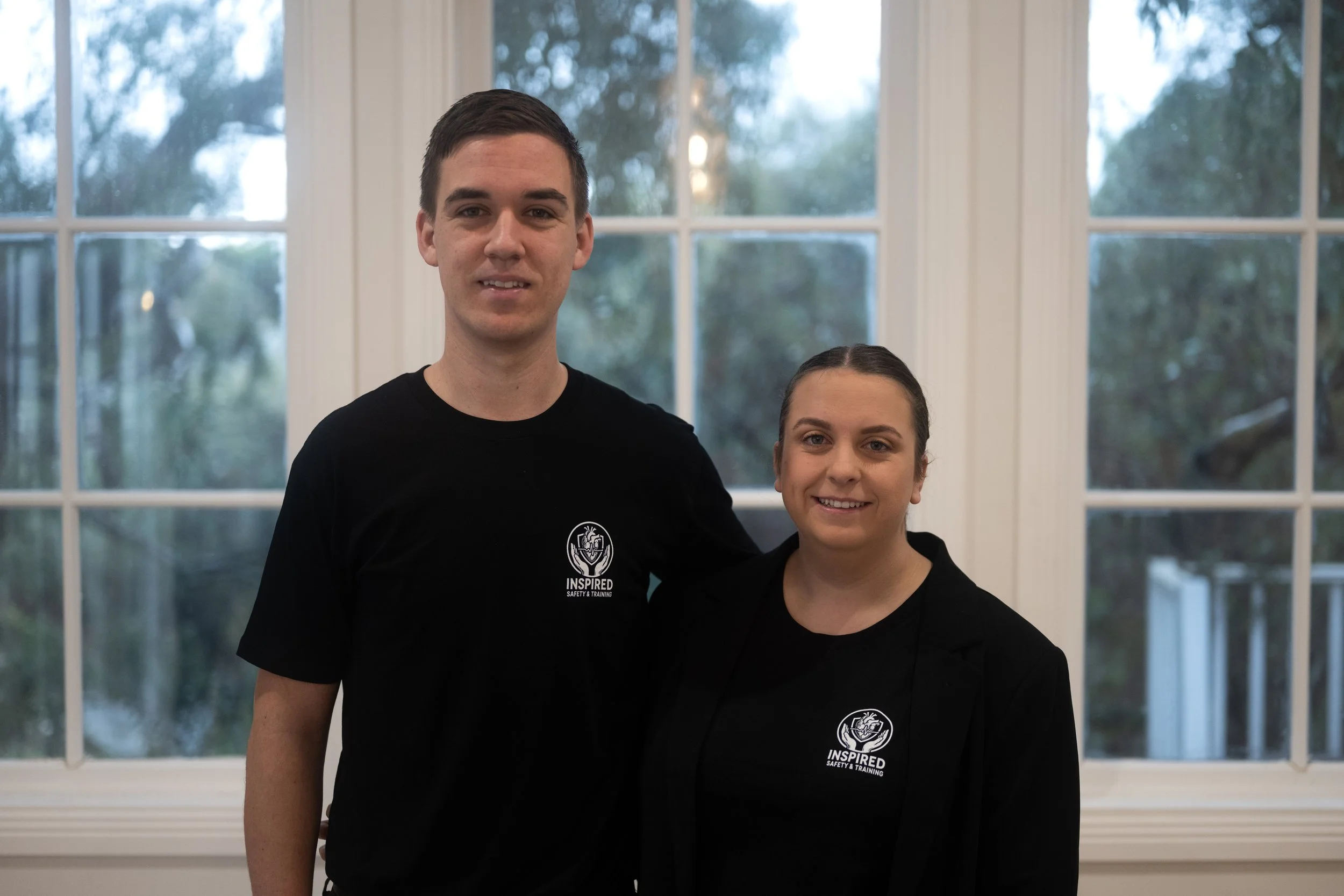 A man and woman standing side by side indoors near a large window, both wearing black shirts with a logo that says 'INSPIRED' and a symbol, smiling at the camera.