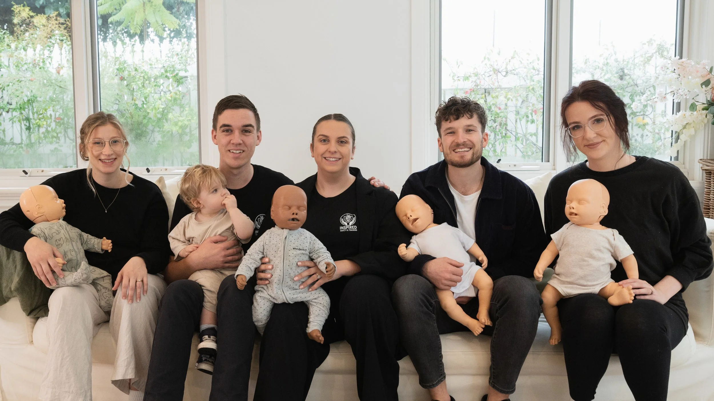 Group of six adults sitting on a couch in a bright room holding baby dolls, with a large window behind them and outdoor greenery visible.