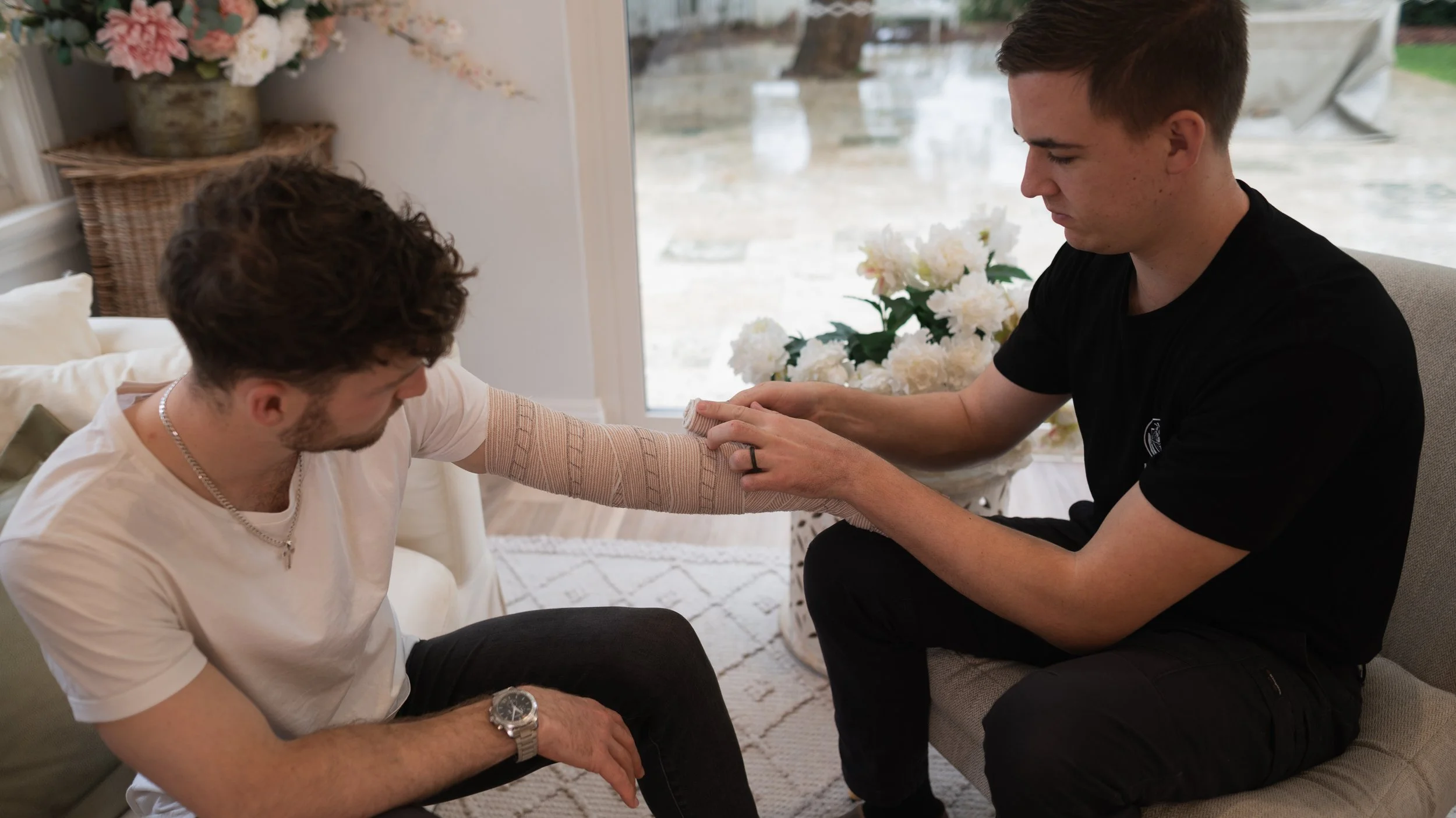 A man with a beard, wearing a white T-shirt, and a silver watch, sitting on a sofa, receiving an arm wrap from another man with short dark hair, wearing a black T-shirt, in a well-lit room with large windows, floral decorations, and a bright interior.