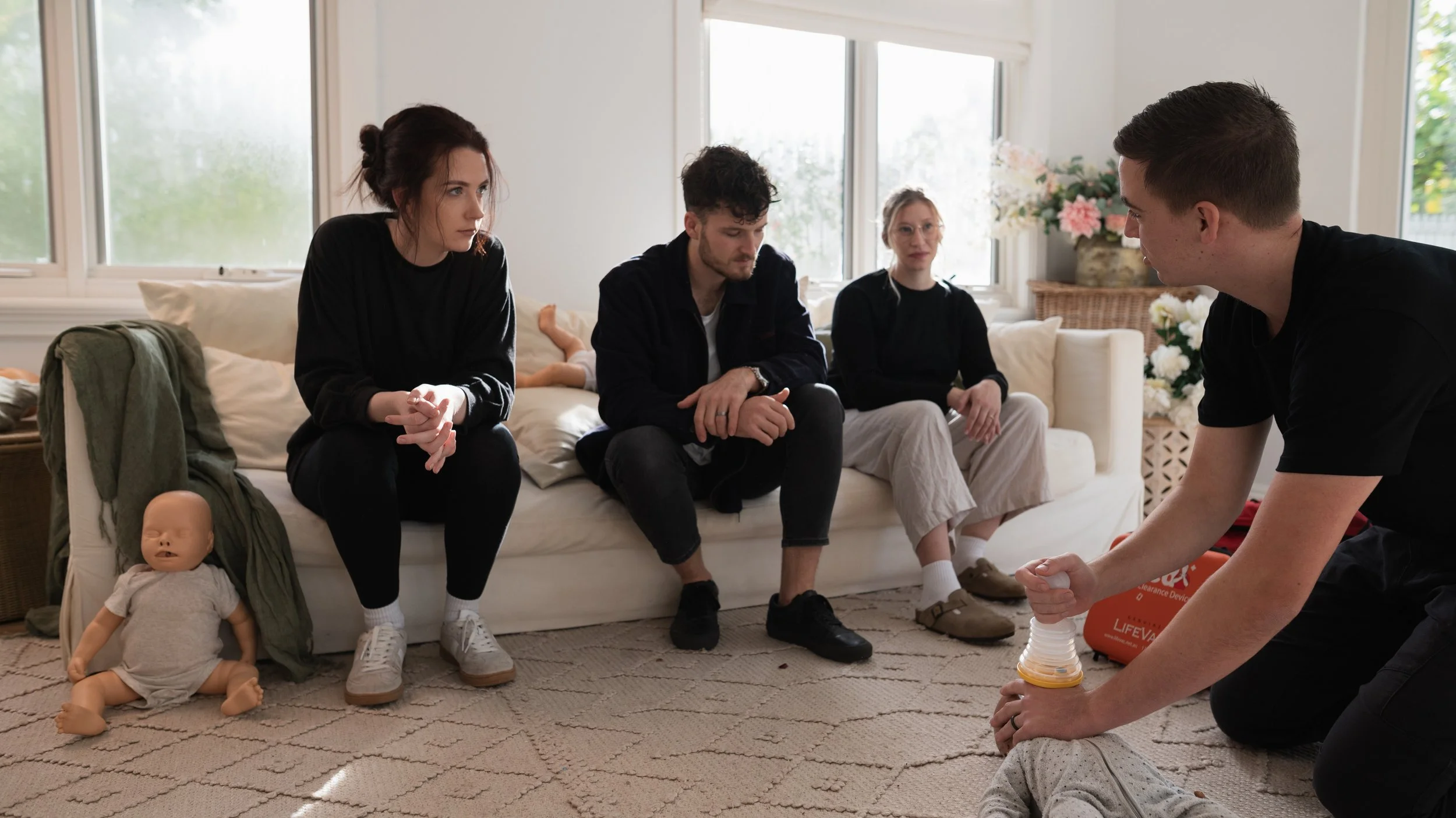 A group of five young adults sitting on a cream-colored couch in a well-lit living room, some with serious expressions, participating in a discussion or activity, with a young man kneeling in front holding a plastic container.