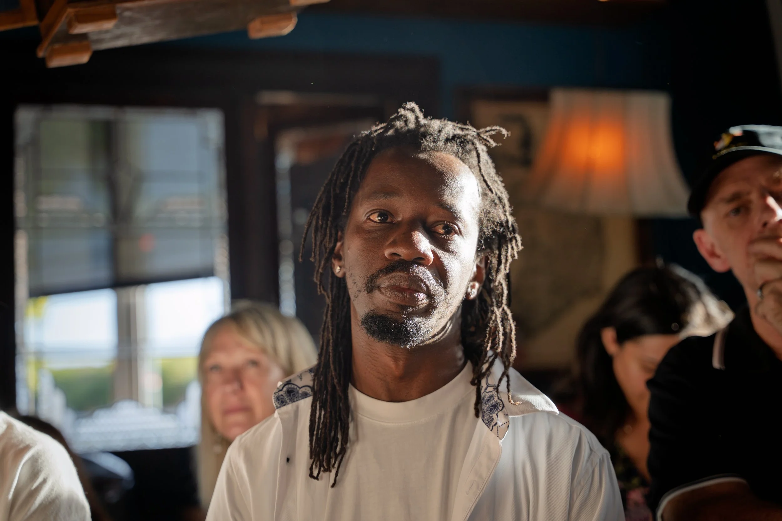 A man with long dreadlocks and earrings, wearing a white shirt, sitting among other people in a dimly lit indoor space with warm lighting and large windows.