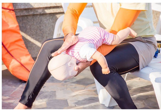 Person performing infant CPR on a mannequin outdoors, with another person assisting.