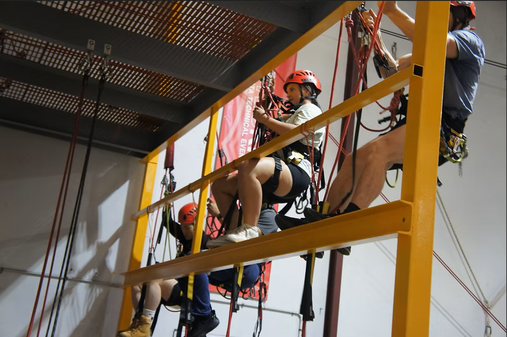 Children and adults wearing helmets and harnesses on a climbing platform with safety gear in an indoor adventure park.