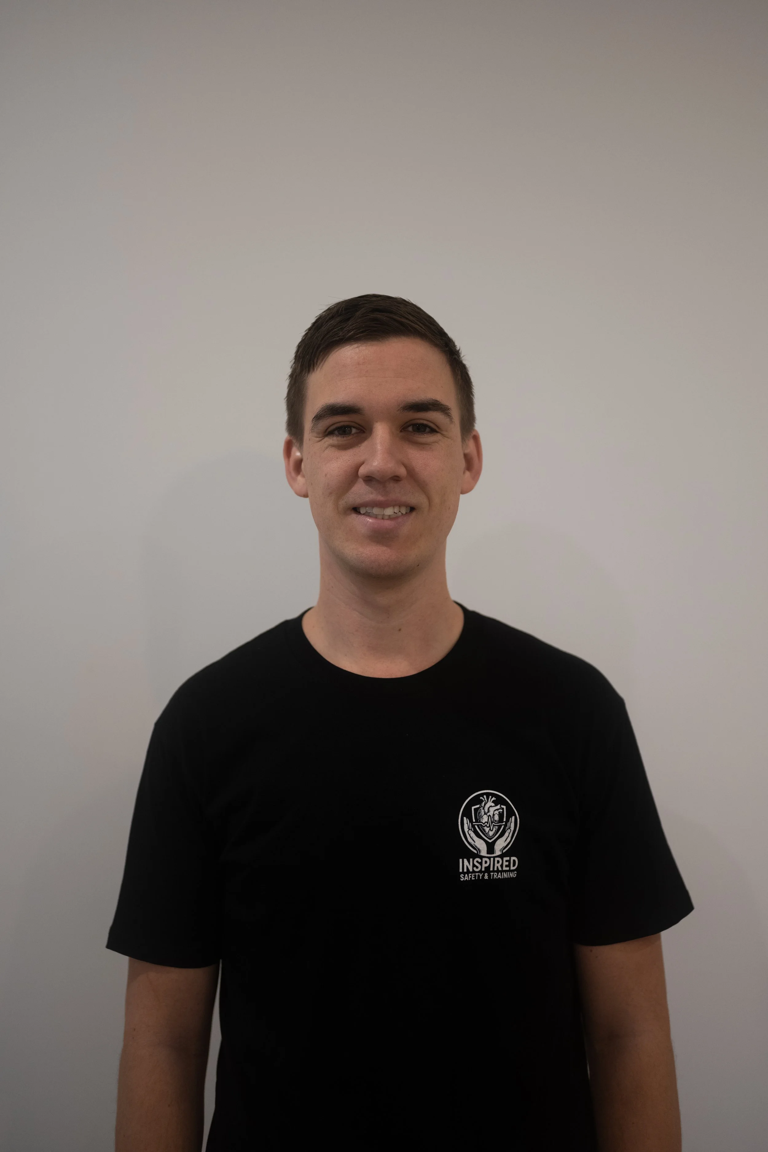 A young man with short dark hair, wearing a black t-shirt with a logo that reads 'Inspired Safety & Training,' standing against a plain white wall.