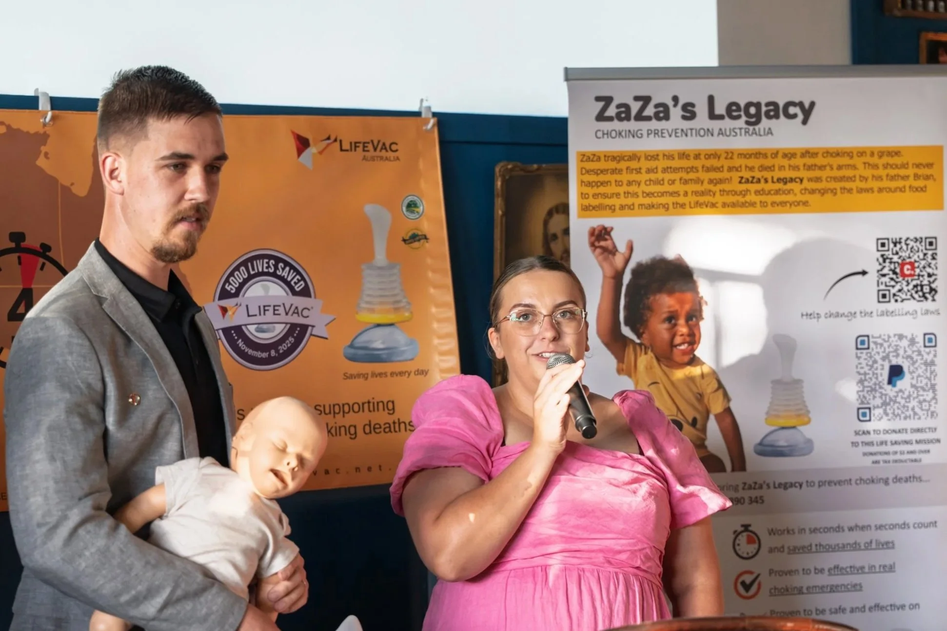 A woman in a pink dress holding a microphone, standing next to a man in a gray blazer, with a young child in her arms, at a presentation about choking prevention by ZaZa's Legacy, with informational posters and banners in the background.