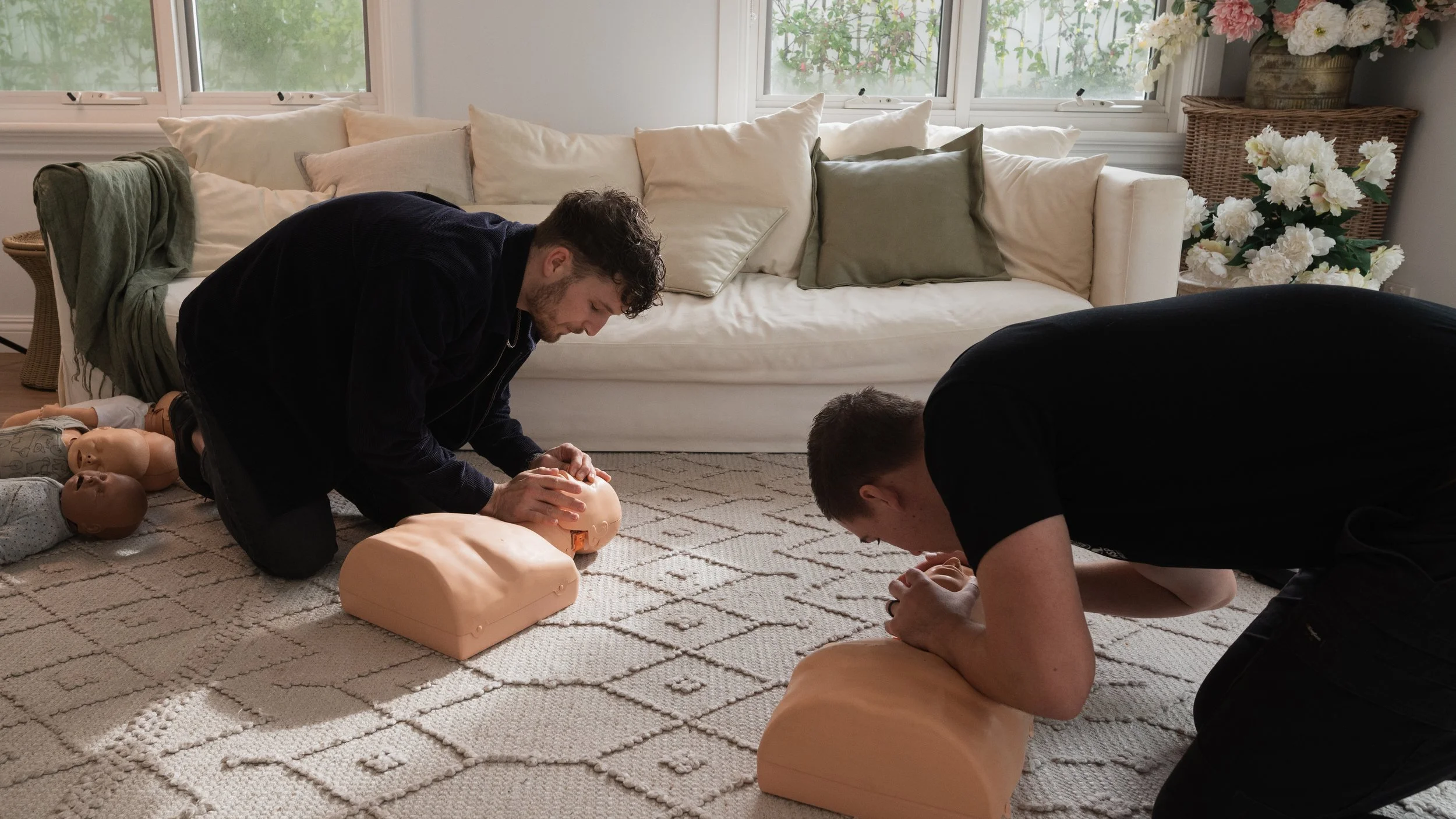 Two people practicing CPR on mannequins in a living room with a white couch and large window.