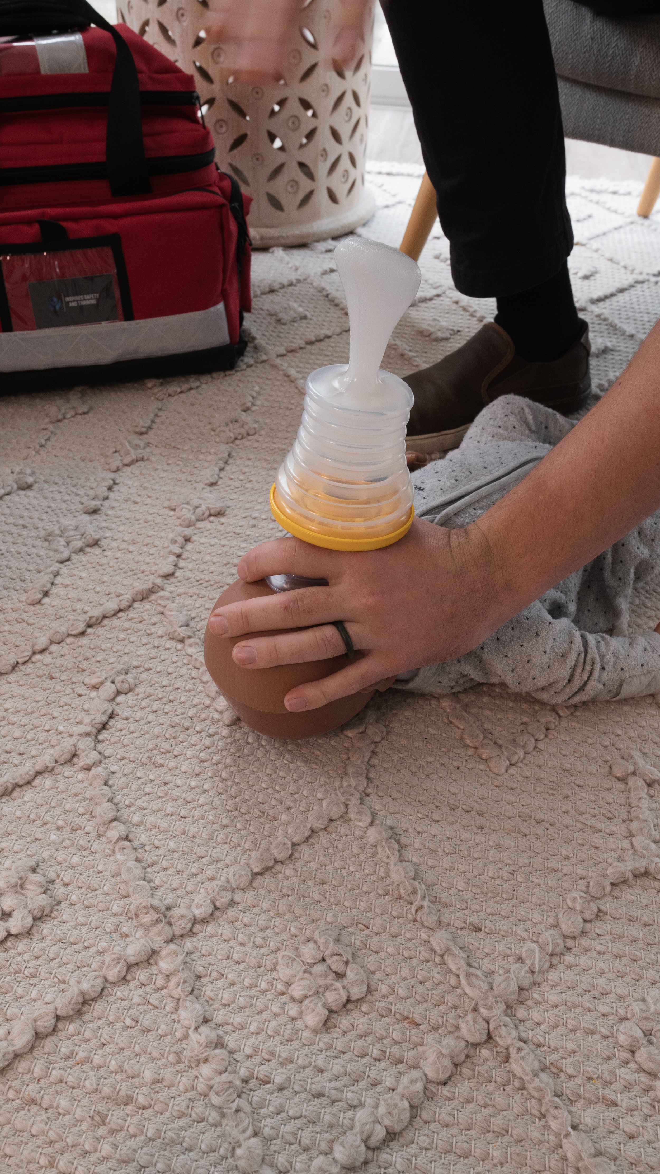 Person holding a baby bottle with a nipple, on a beige textured rug, with a red bag and patterned cloth in the background.