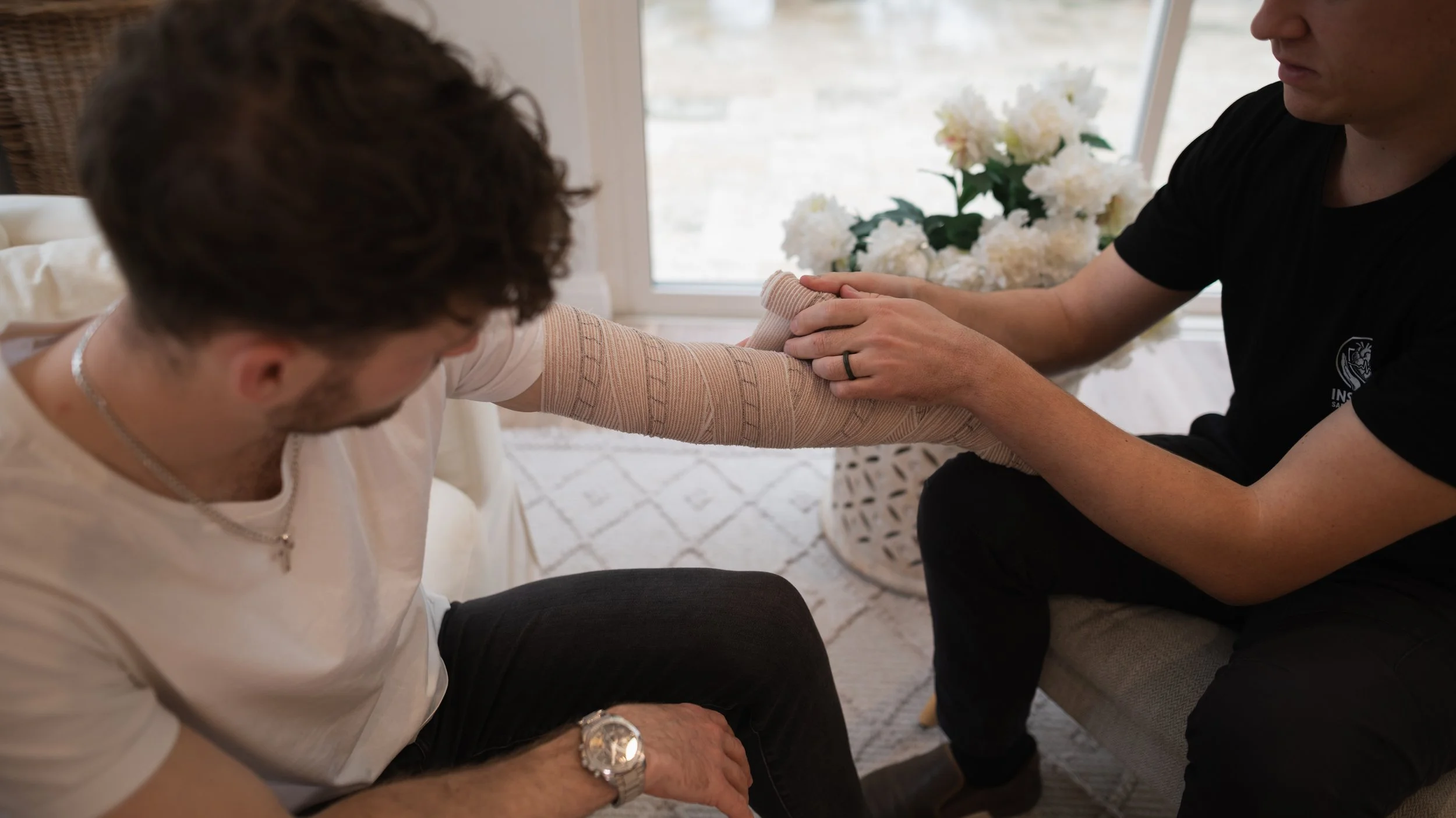 A person receiving a medical treatment on their arm from a healthcare professional inside a bright room with white flowers in the background.