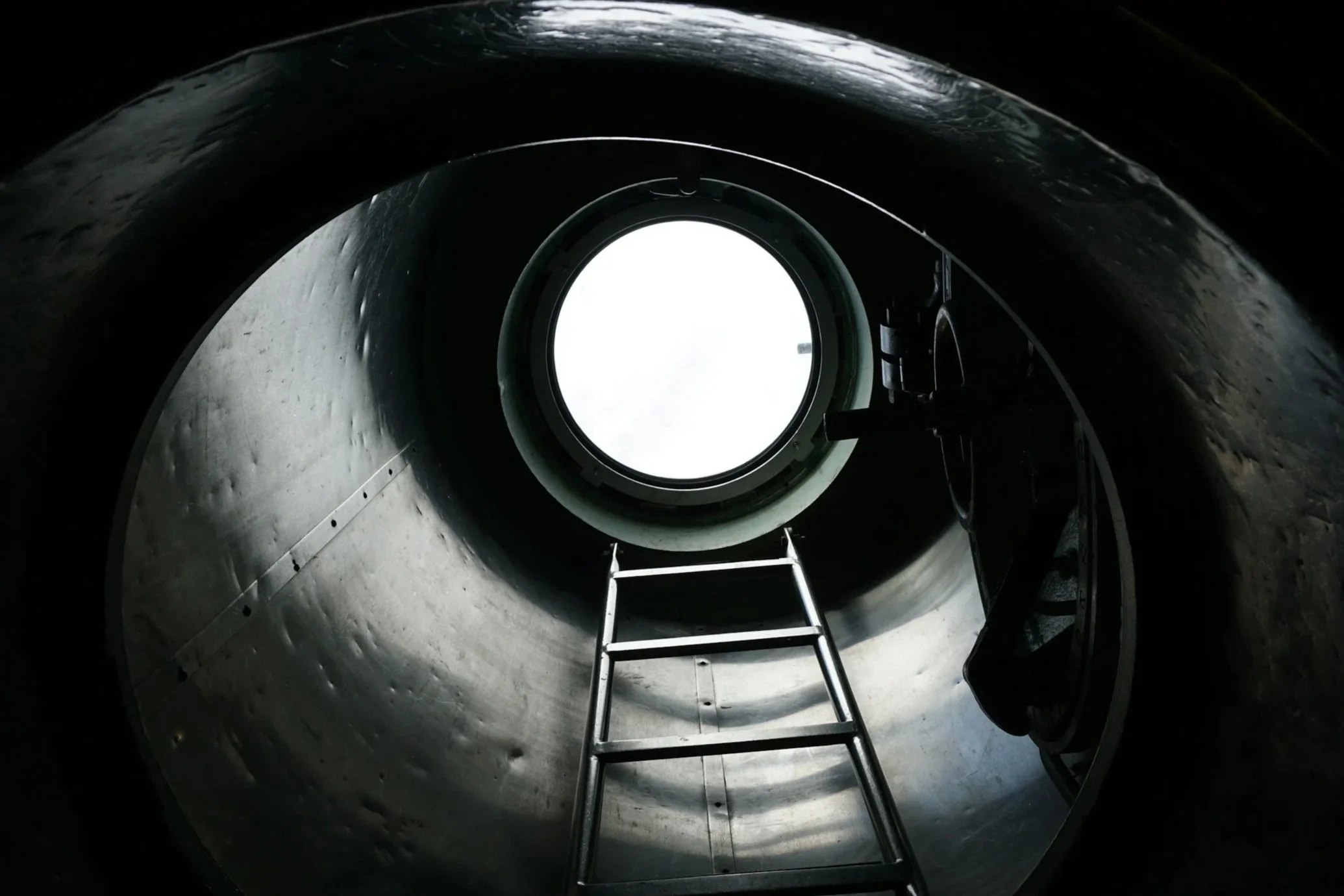 The interior of a metallic rocket or spacecraft viewed from inside with a large circular opening in the wall, a ladder leading up to the opening, and some equipment attached to the wall.