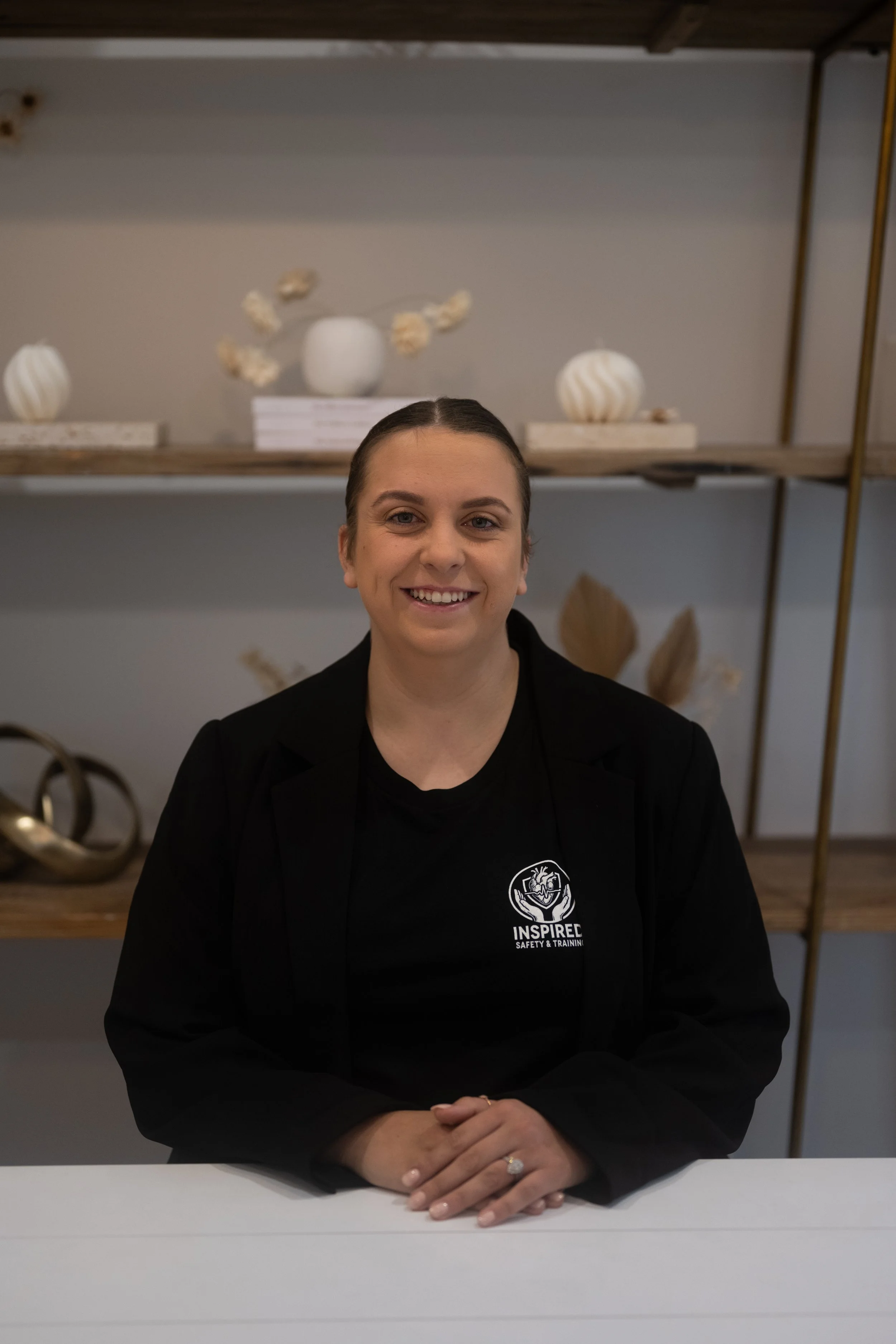 A woman smiling, sitting at a table with her hands clasped, wearing a black blazer and a black T-shirt with a logo that says "INSPIRE SAFETY & TRAINING." In the background are decorative objects on shelves, including white vases and sculptures.
