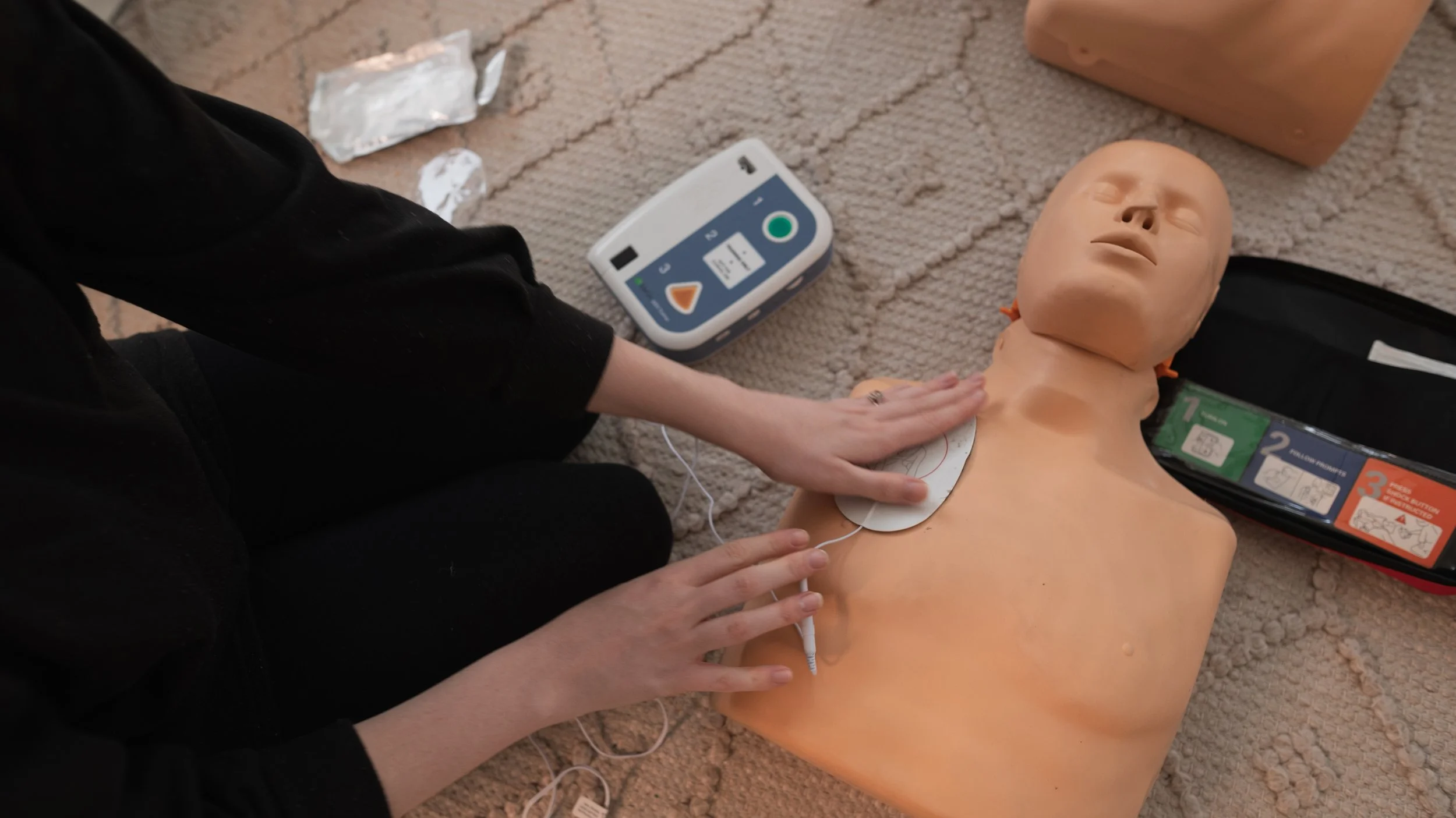 A person performing CPR on a medical training mannequin lying on a beige carpet. The person's hands are on the mannequin's chest, and there is an automated external defibrillator (AED) nearby.