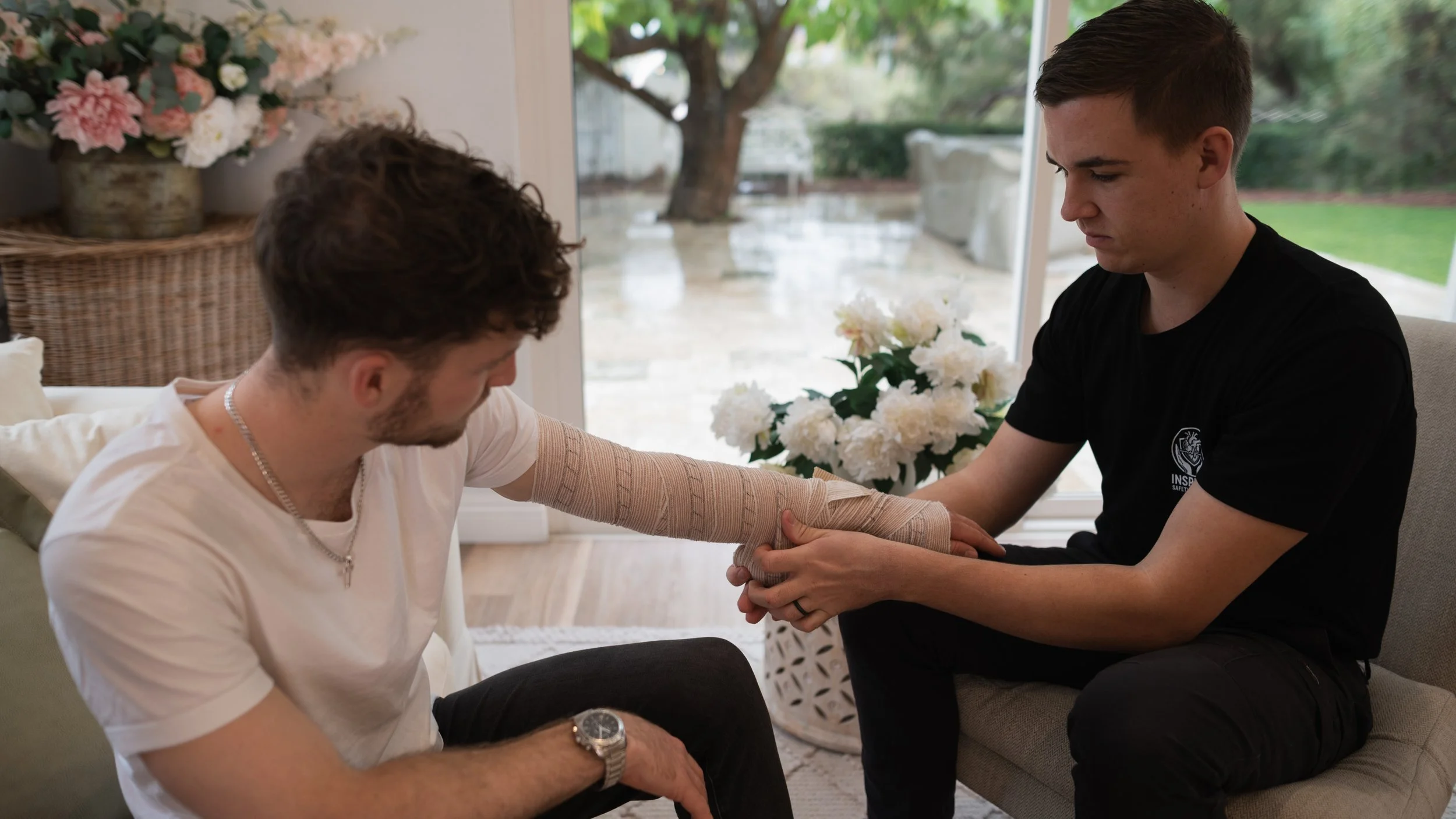A young man with short dark hair and a young woman with curly brown hair sitting indoors near a large window, with a tree and greenery visible outside. The young man is helping the woman by wrapping her arm in a bandage, and she is wearing a white t-shirt and a watch on her left wrist. The woman is seated, and the man is kneeling beside her, focused on tending to her arm. There are flowers in a pot on the windowsill.