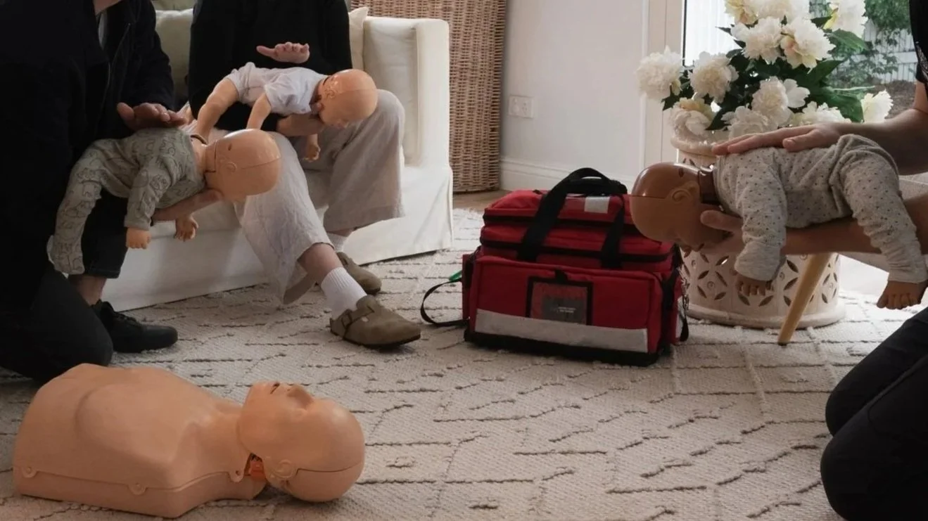 People practicing infant CPR on mannequins in a home setting with a red emergency medical bag nearby.