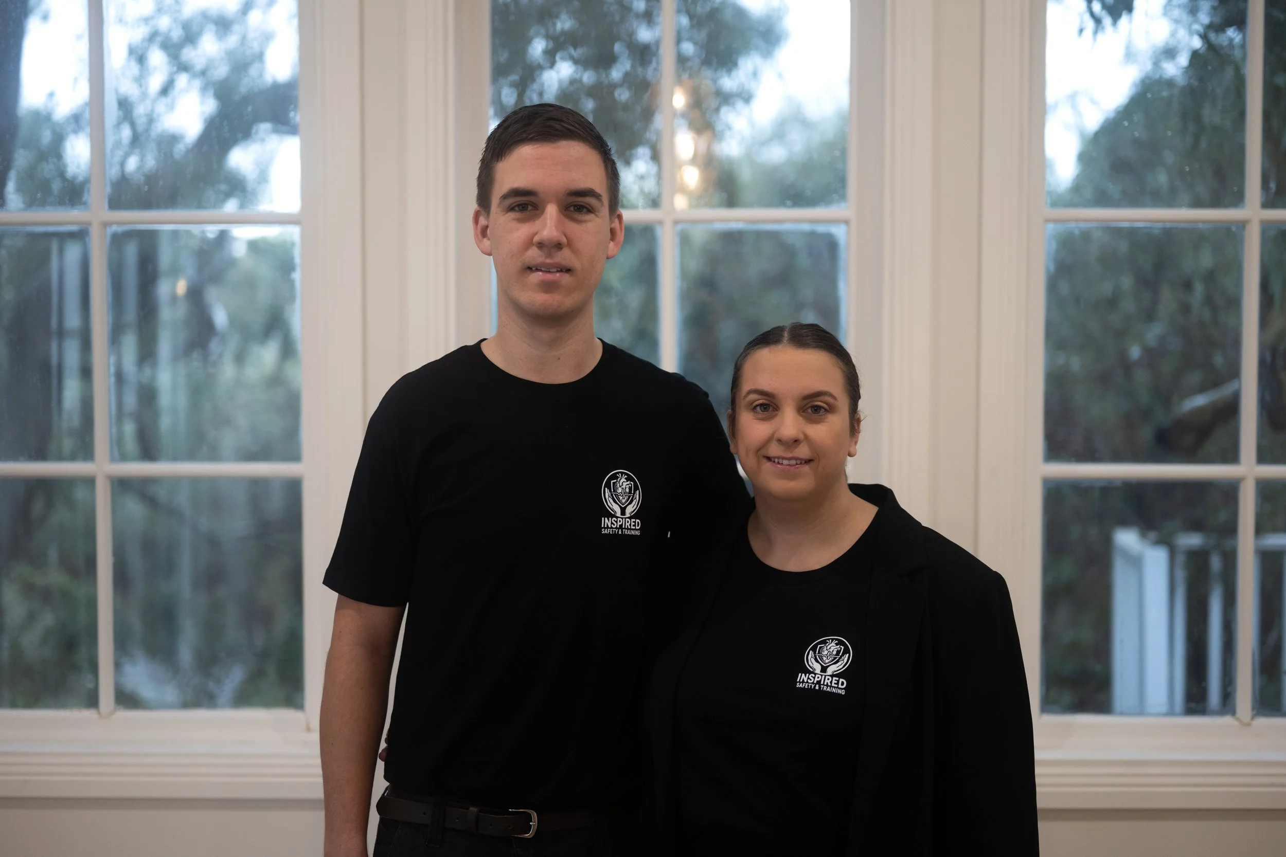 A man and woman standing side by side indoors in front of large windows, both wearing black shirts with a logo and text that says 'Inspired Safety & Training'.