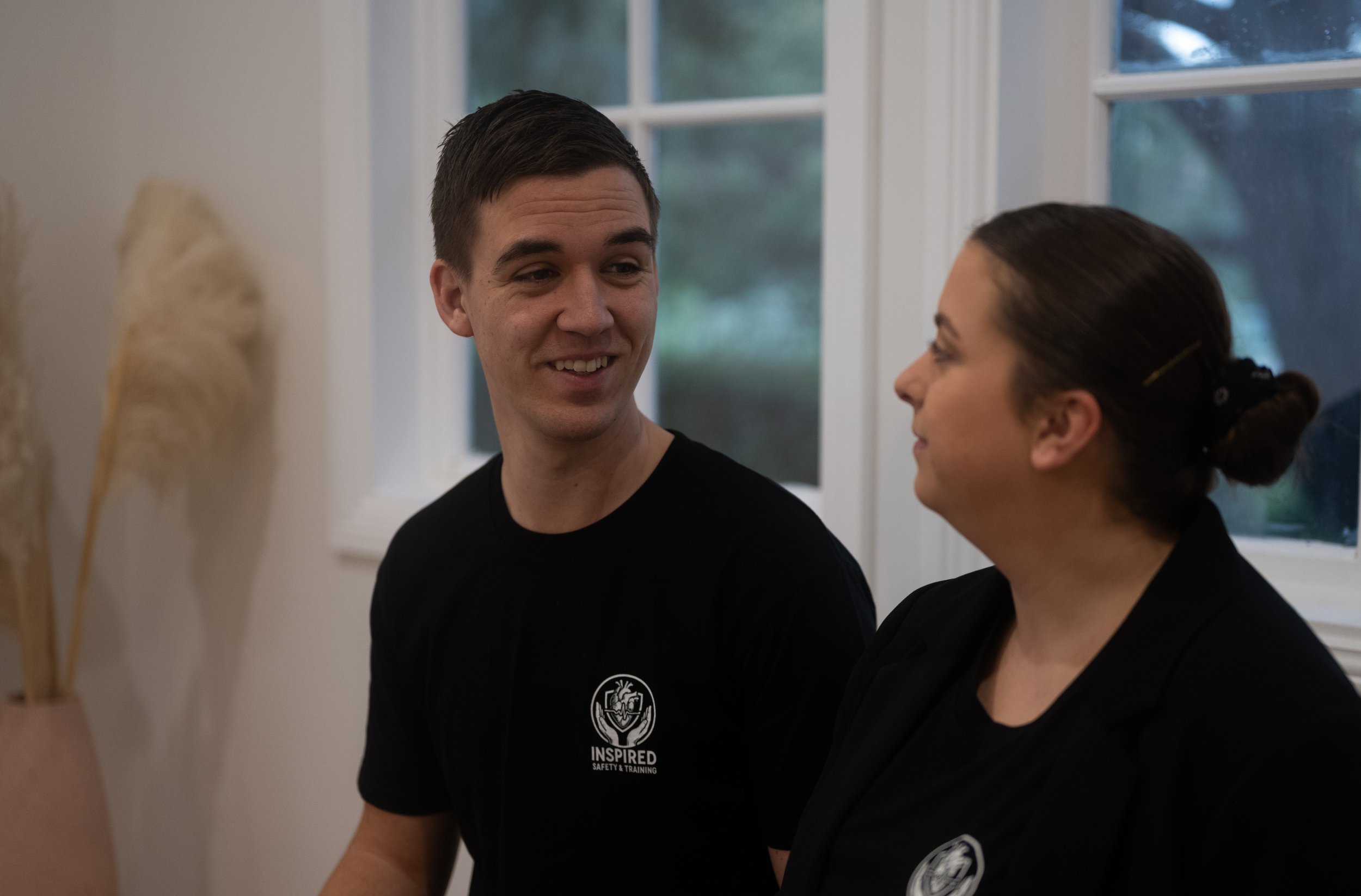 A young man and a woman are standing close to each other indoors, smiling and looking at each other. They are both wearing black shirts, and the woman's shirt has a logo that says "INSPIRED SAFETY & TRAINING." There are large windows behind them, showing a green outdoor view, and a blurred sculpture or decorative object is partially visible on the left side of the image.