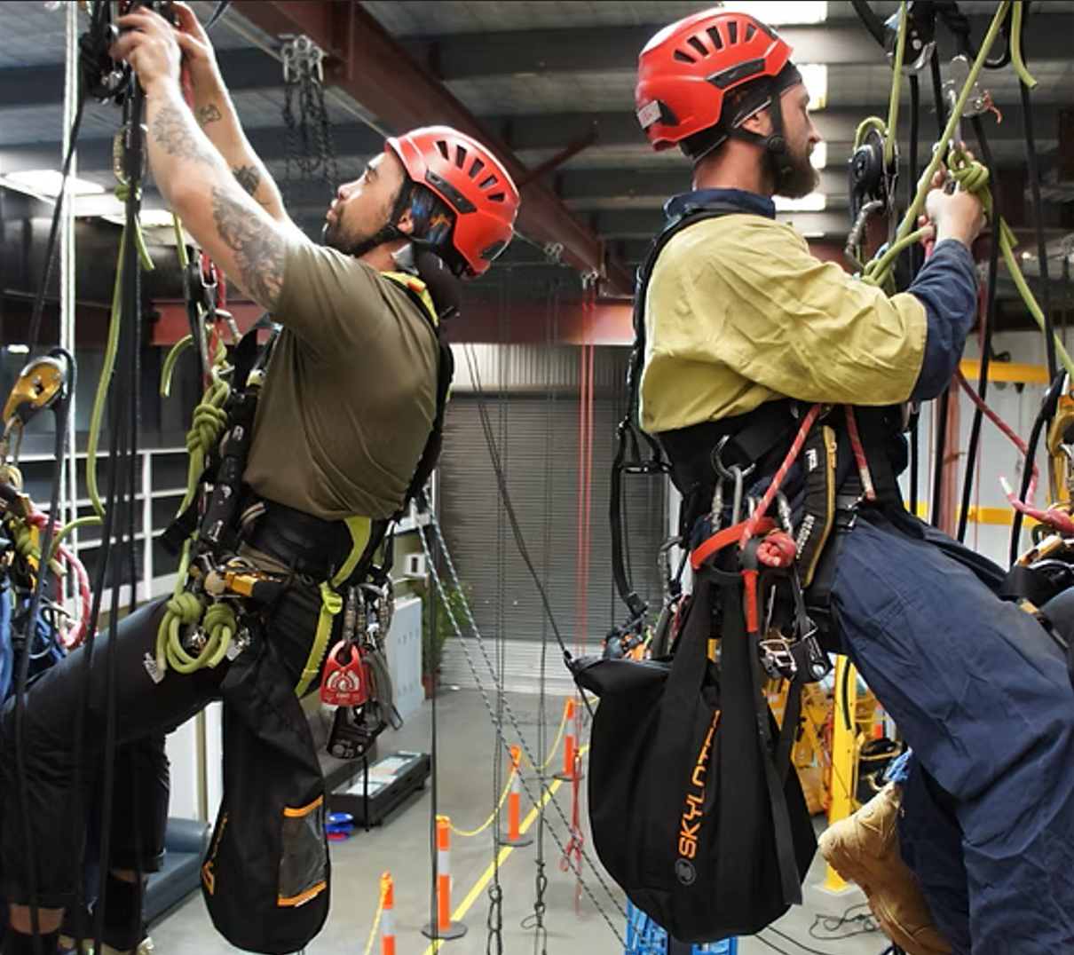 Two men with red helmets climbing harnesses are preparing for a ropes course or high ropes activity indoors, with safety gear and equipment around them.