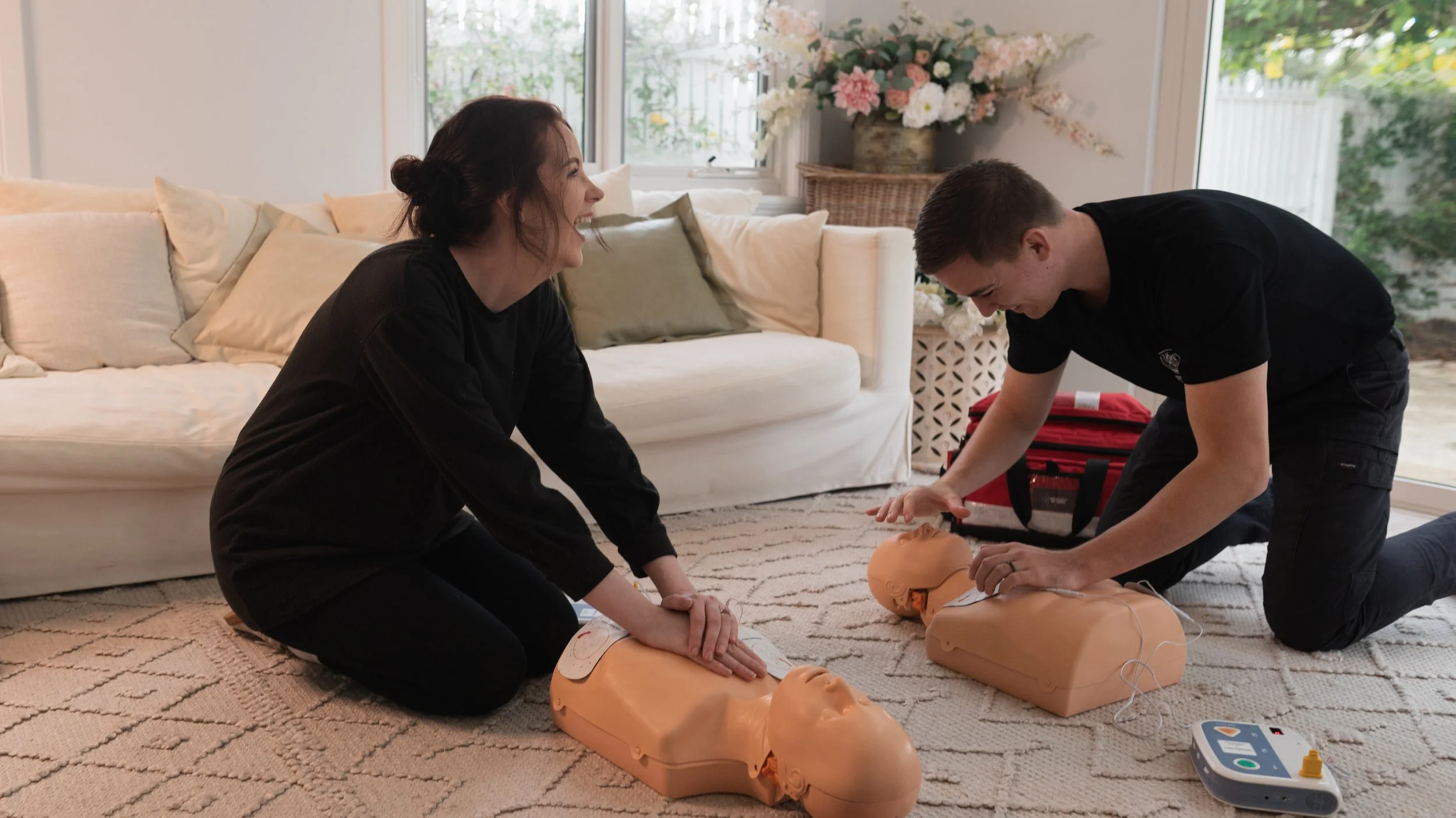 Two people performing CPR on a training mannequin in a living room with a cream-colored couch and a large floral arrangement in the background.