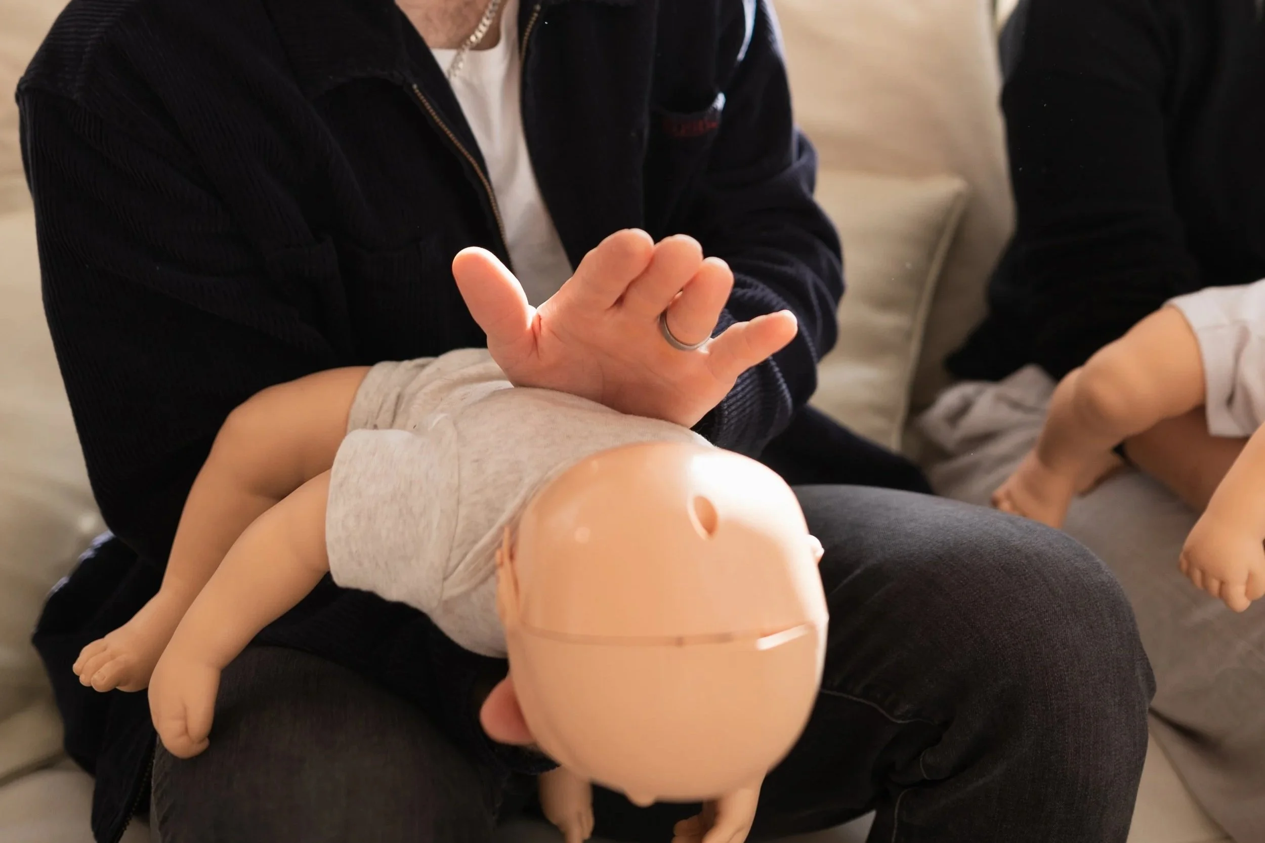 A person practicing CPR on a mannequin baby, with another person watching. The instructor is demonstrating a breathing technique with a gloved hand.
