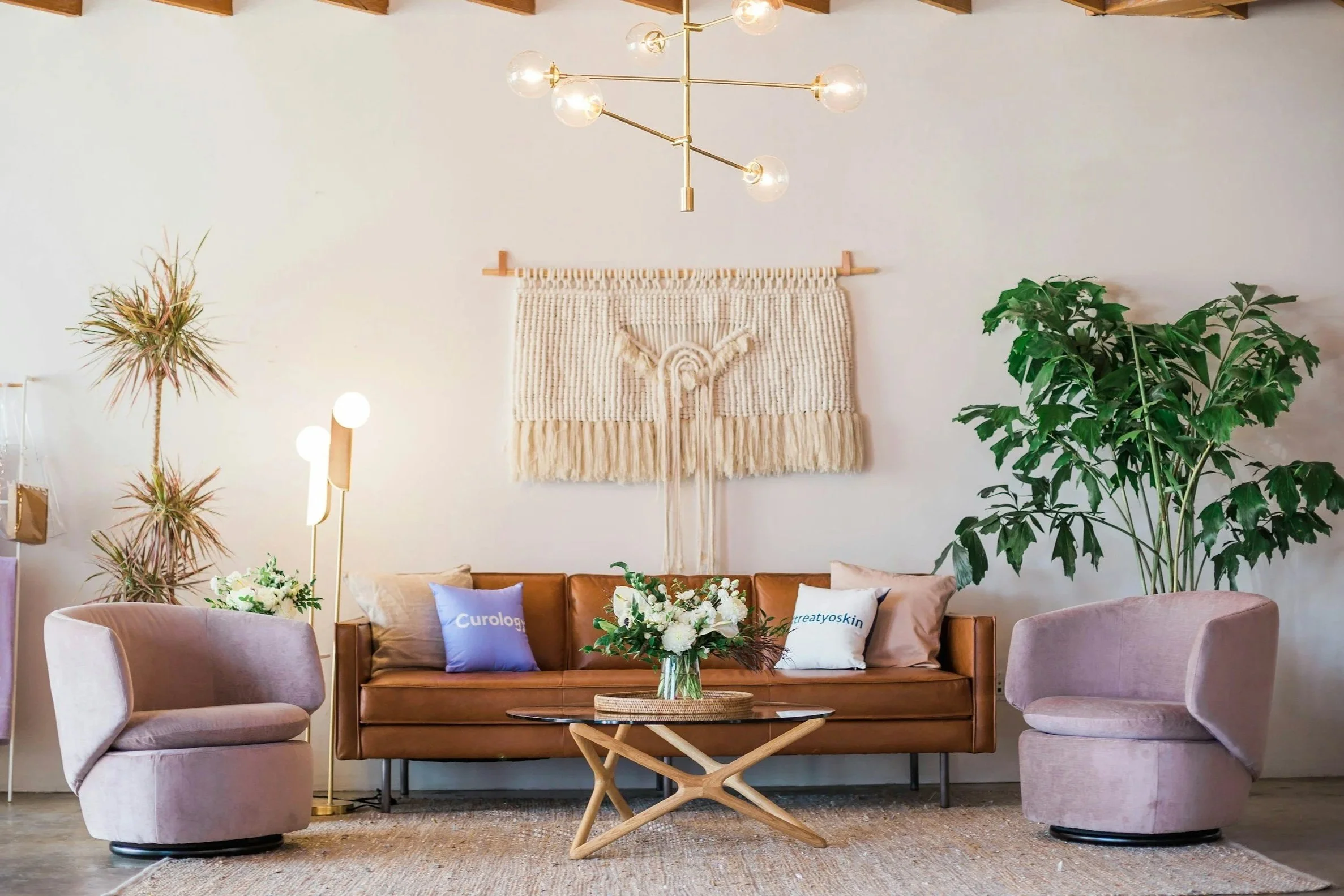 A modern living room with a tan leather sofa flanked by two pink upholstered swivel chairs. A wooden coffee table with a glass top features a floral arrangement. Two decorative pillows on the sofa A large green plant is on the right, and a smaller plant on the left. A textured wall hanging with fringe is on the white wall above the sofa. A contemporary chandelier with multiple exposed bulbs hangs from the ceiling.