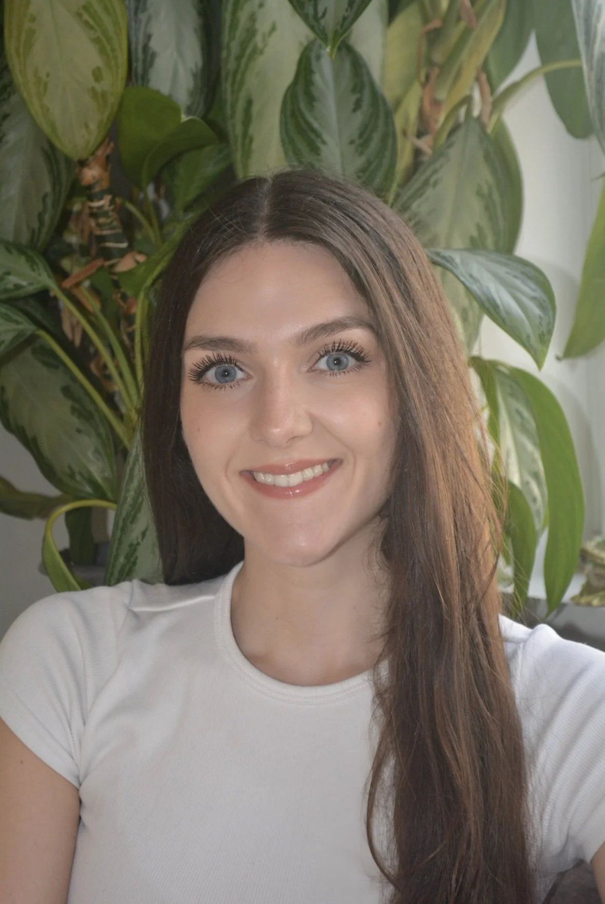 A young woman with long brown hair and blue eyes smiling in front of a large green leafy plant.