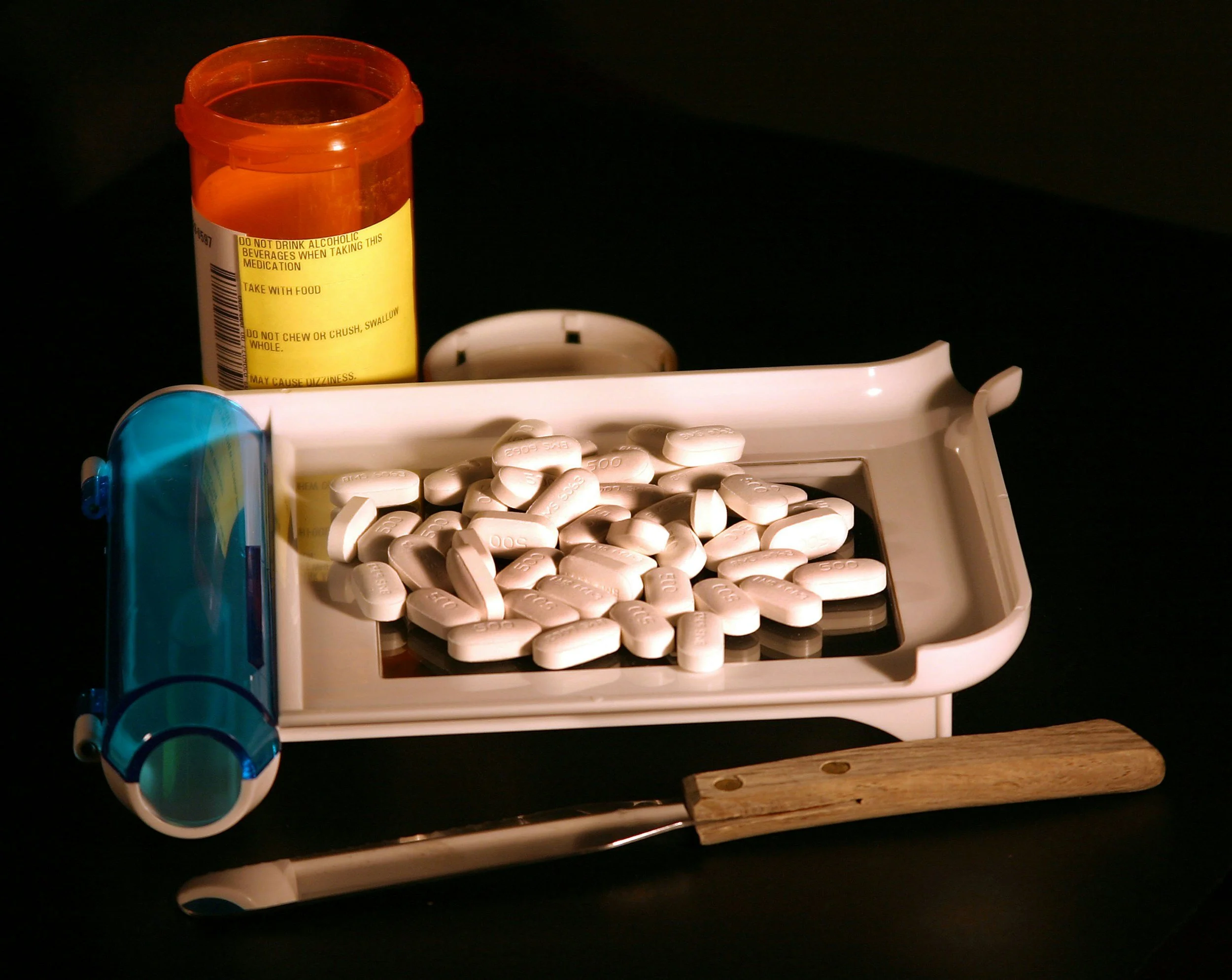 A pill counting tray with white tablets, a prescription bottle with a yellow label, a blue and white prescription bottle holder, and a pair of tweezers on a dark surface.