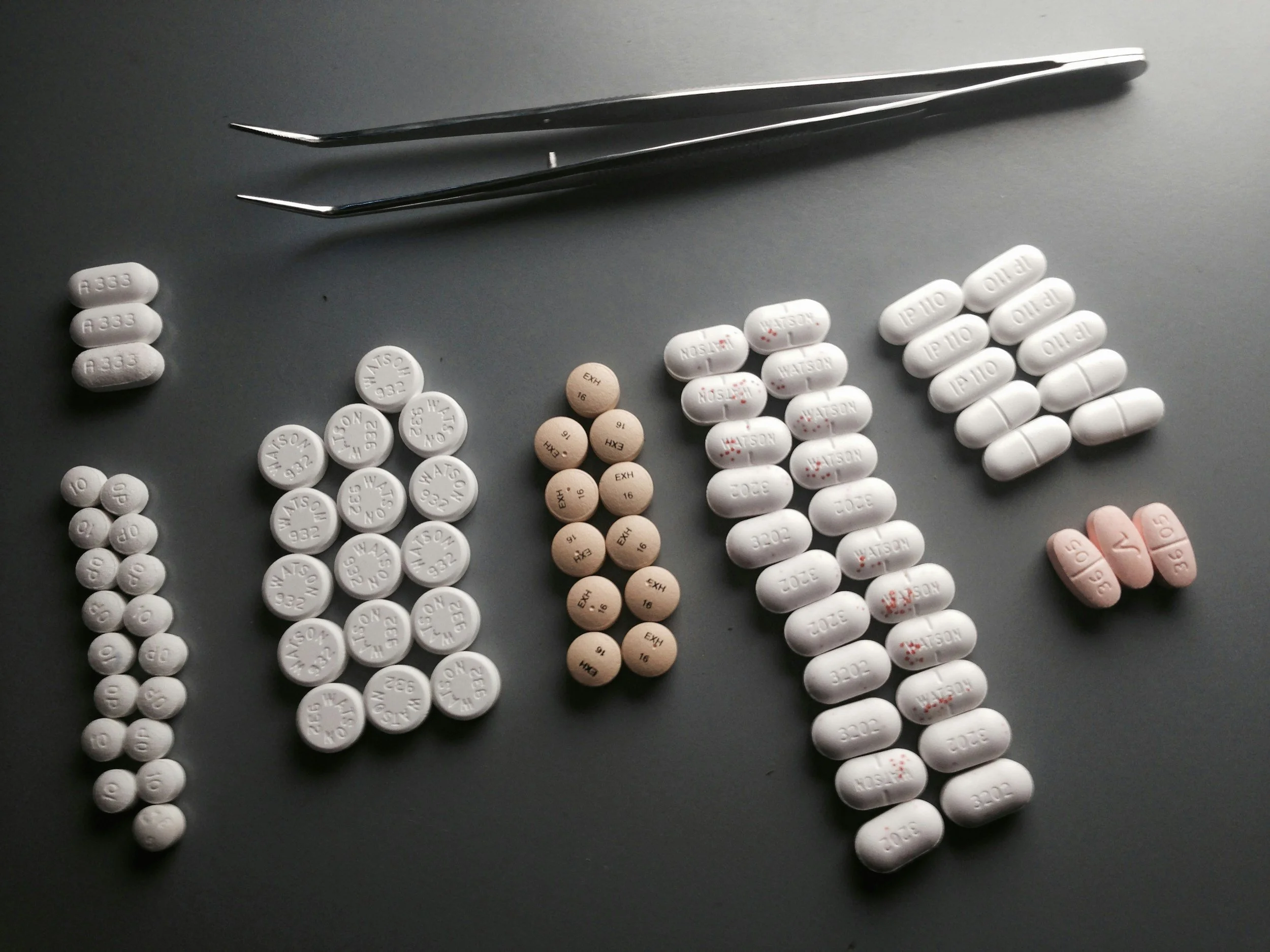 An assortment of prescription pills and tablets arranged on a gray surface with a pair of tweezers at the top.