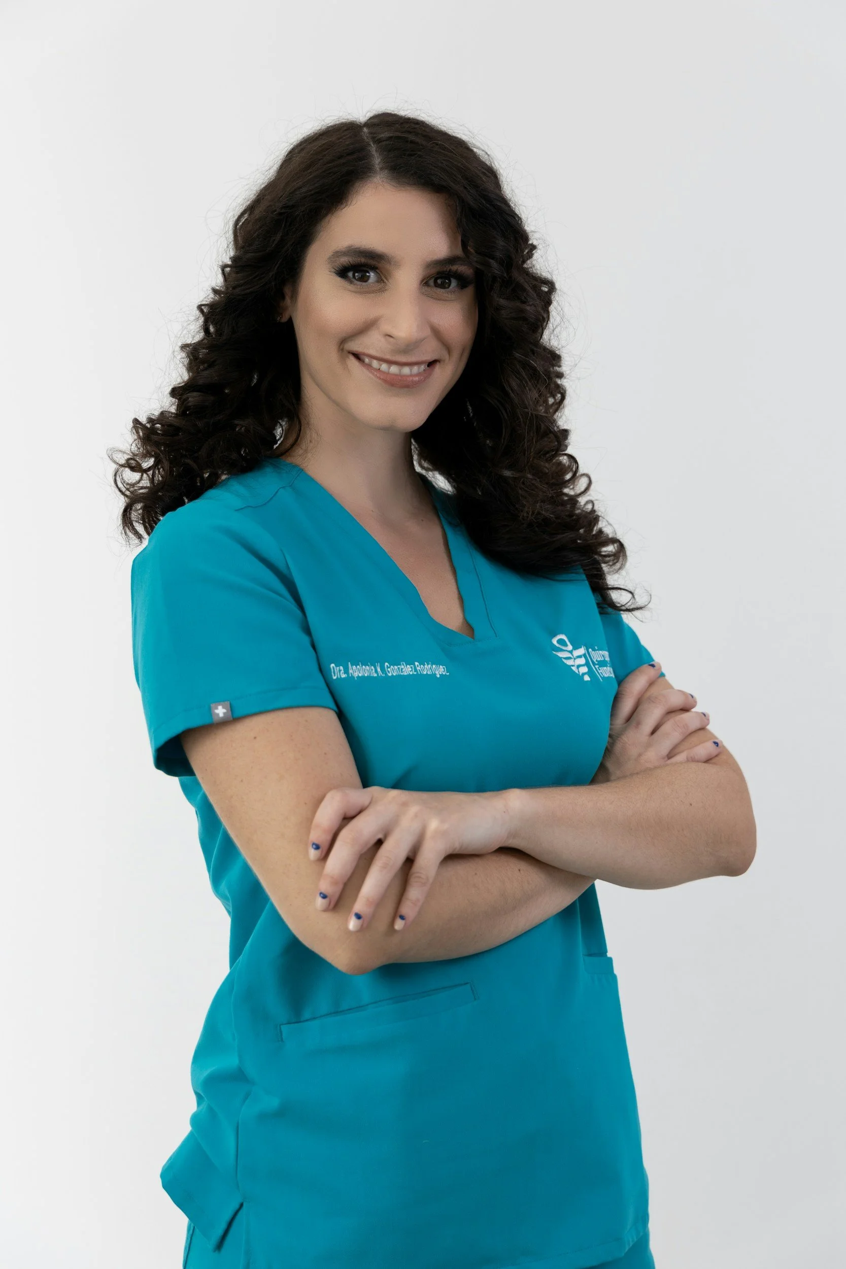 A woman in teal medical scrubs with the name tag and the logo of a healthcare facility, smiling with arms crossed, standing against a white background.